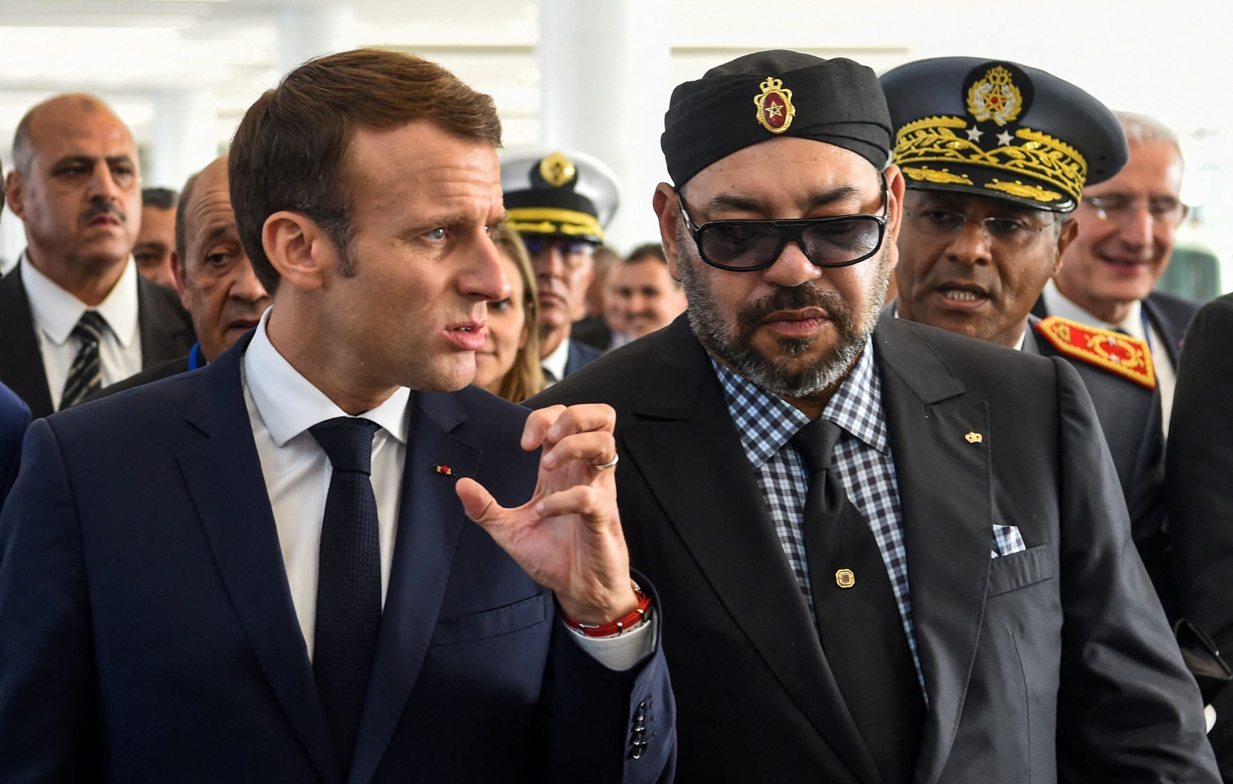 L'image montre deux hommes en discussion, entourés de plusieurs personnes. L'un porte un costume sombre et une cravate, tandis que l'autre est habillé d'une manière plus formelle, avec des lunettes de soleil et un turban. Ils semblent engagés dans une conversation sérieuse, et l'environnement semble être une cérémonie officielle ou un événement important. Les expressions sur leurs visages suggèrent qu'ils sont en train de partager des idées ou des informations.