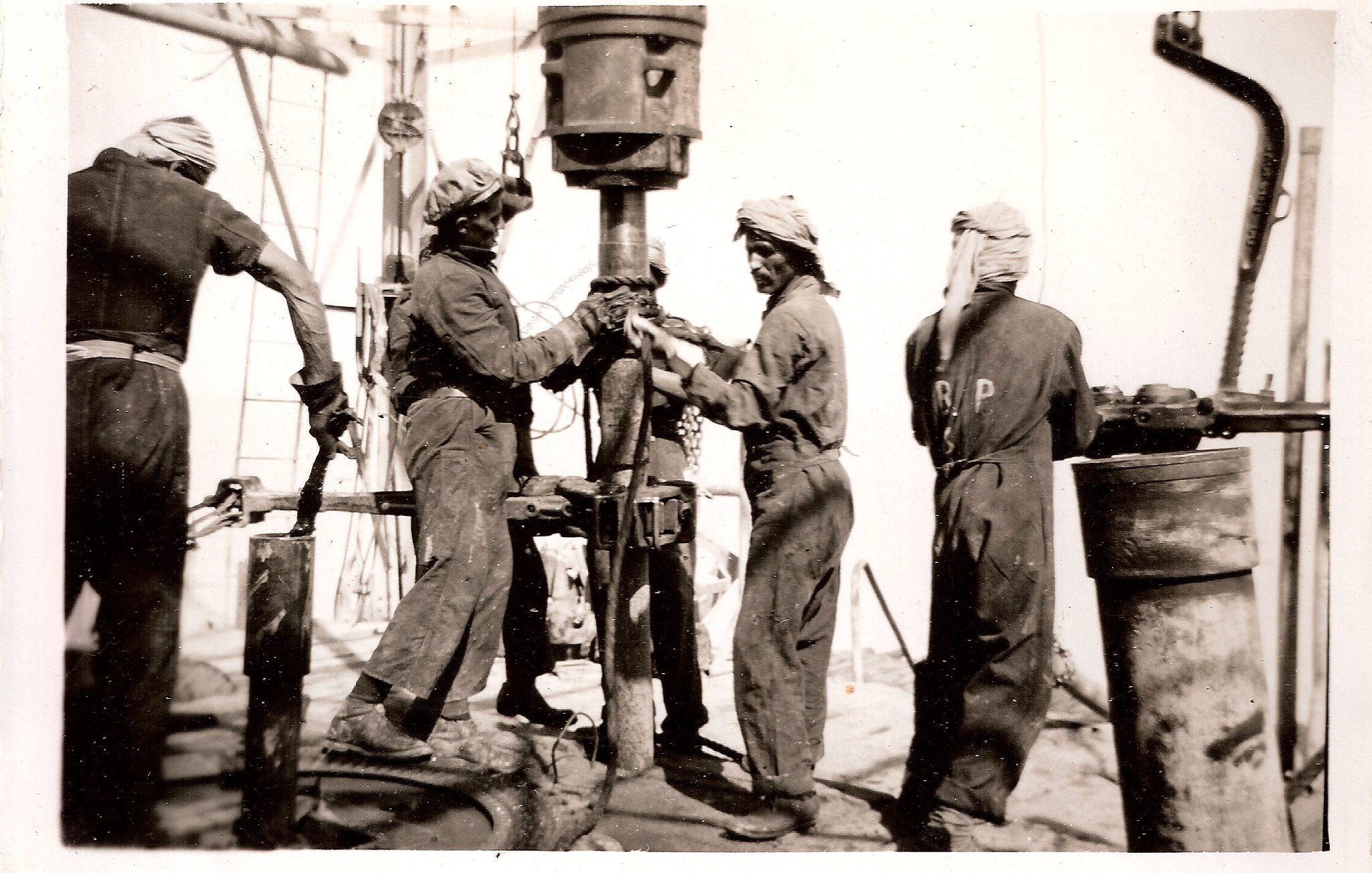 L'image représente un groupe de travailleurs en train de manipuler un équipement de forage, vraisemblablement sur un chantier pétrolier ou minier. Les ouvriers portent des vêtements de travail et des turbans, probablement pour se protéger de la poussière ou de la chaleur. Ils semblent concentrés sur leur tâche, travaillant ensemble sur une machine visible au centre de l'image. L'ambiance paraît industrialisée et intense, typique des travaux de forage au début du XXe siècle.