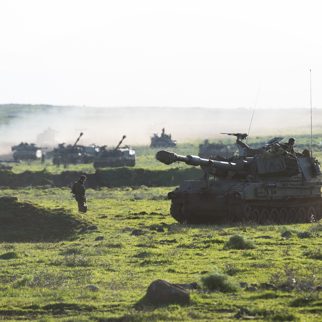 L'image montre un paysage militaire verdoyant avec des chars d'assaut et d'autres véhicules en arrière-plan. On peut voir un char principal en premier plan, déployé sur un terrain herbeux, tandis que plusieurs autres véhicules sont visibles plus loin, entourés d'une légère brume ou fumée. Quelques soldats sont présents, donnant une impression d'activité militaire dans un environnement naturel. La lumière est douce, suggérant peut-être un moment du jour comme le lever ou le coucher du soleil.