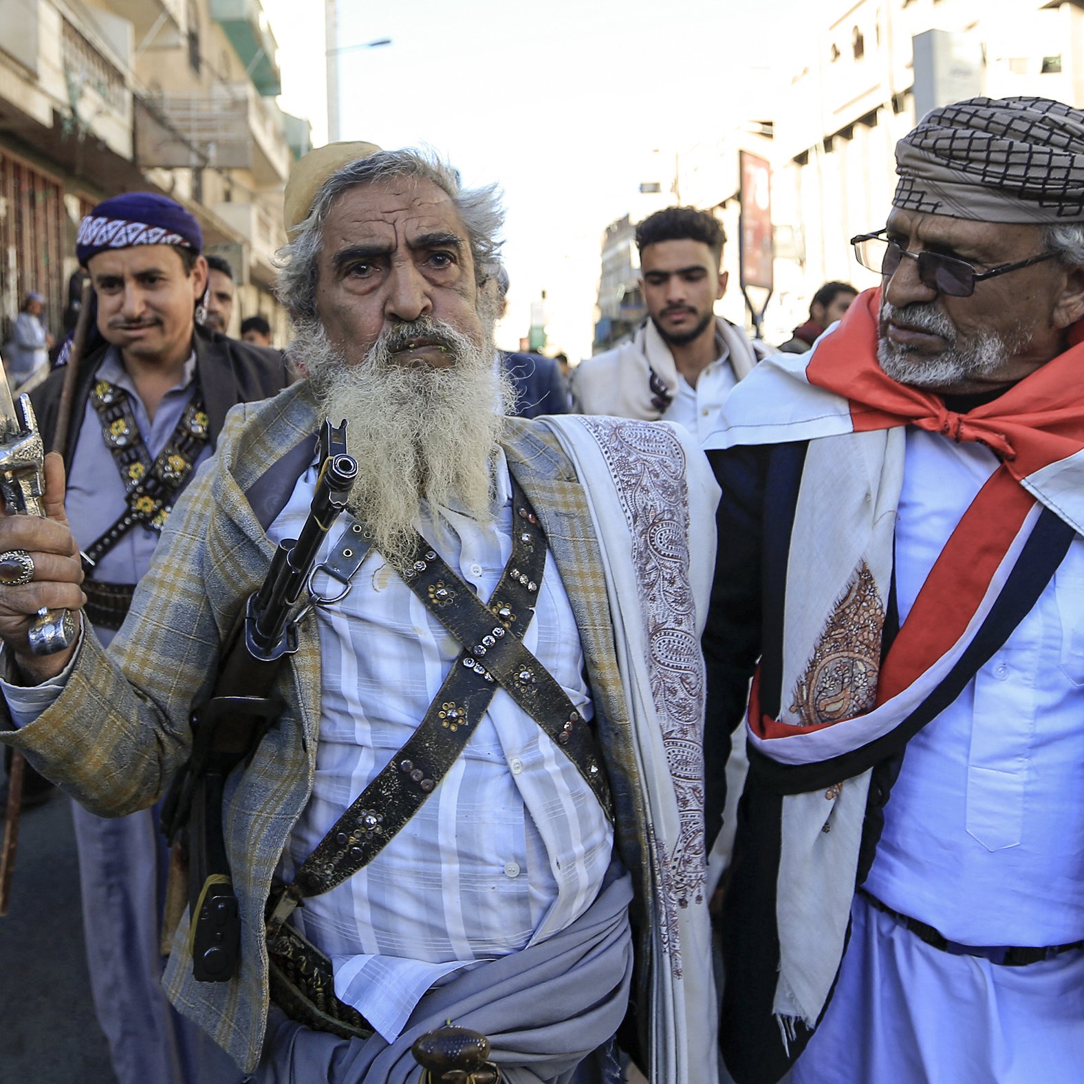 در این تصویر، چند مرد در حال راه رفتن در یک خیابان هستند. یکی از آن‌ها، که در پیش‌زمینه قرار دارد، دمی بلند و ریش سفیدی دارد و یک شمشیر در دست دارد. او لباس سنتی یمنی به تن کرده و به نظر می‌رسد که مقام یا اهمیت خاصی دارد. دیگر مردان نیز لباس‌های سنتی بر تن دارند و برخی از آن‌ها سلاح‌هایی در دست دارند. جو در تصویر به نظر مهیج و پر از نشاط است و نشان‌دهنده یک گردهمایی یا تجمع اجتماعی می‌باشد.