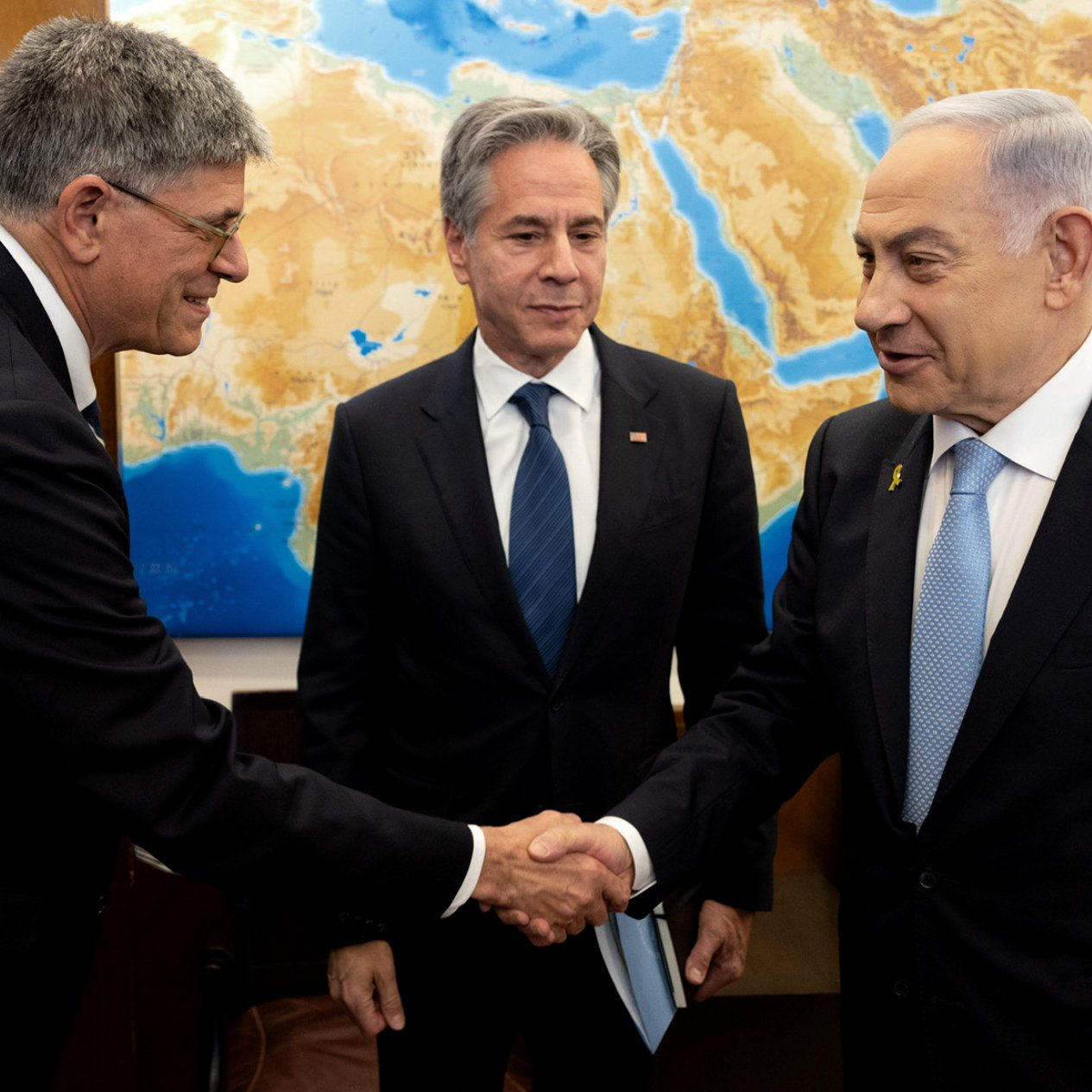 L'image montre trois hommes dans un cadre formel. À gauche, un homme tend la main pour saluer un autre homme à droite, qui semble être le Premier ministre israélien. Au centre, un homme observe la scène. Le fond présente une carte de la région méditerranéenne, ce qui suggère un contexte diplomatique ou de négociations. Les hommes sont habillés de façon professionnelle, ce qui renforce l'atmosphère sérieuse de la rencontre.