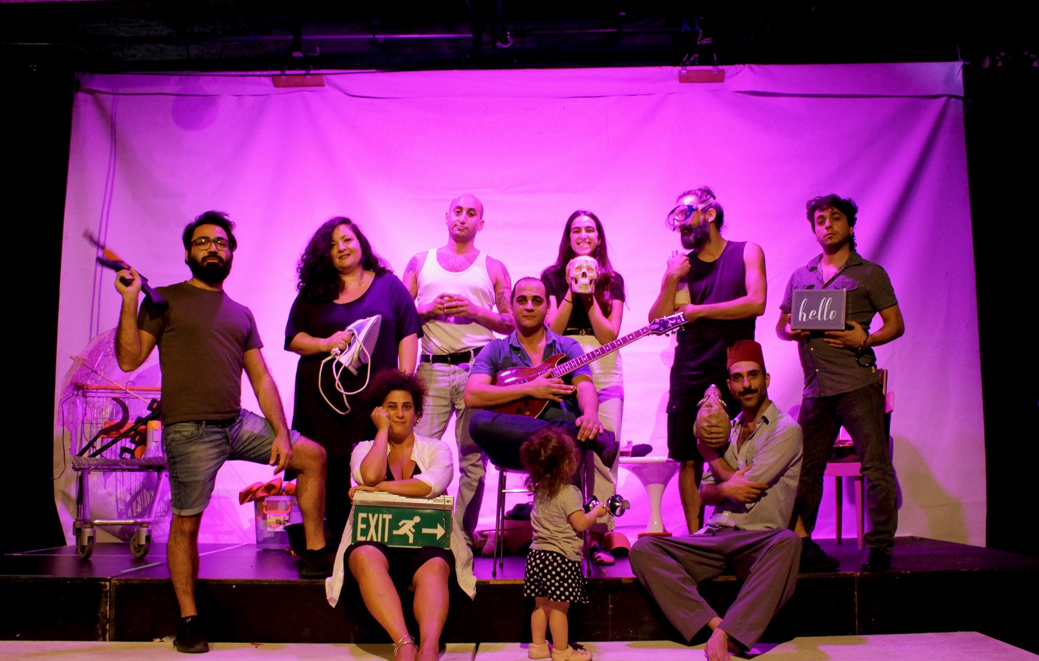L'image montre un groupe de personnes posant sur scène dans une ambiance colorée, avec un éclairage rose. Chaque membre a une pose ou un accessoire distinct, créant un effet visuel dynamique. Certains tiennent des objets variés, comme un instrument de musique, un panneau de sortie et un appareil électronique. Au premier plan, on peut voir une petite fille debout, ajoutant une touche de légèreté à la scène. L'ensemble dégage une atmosphère artistique et joyeuse.