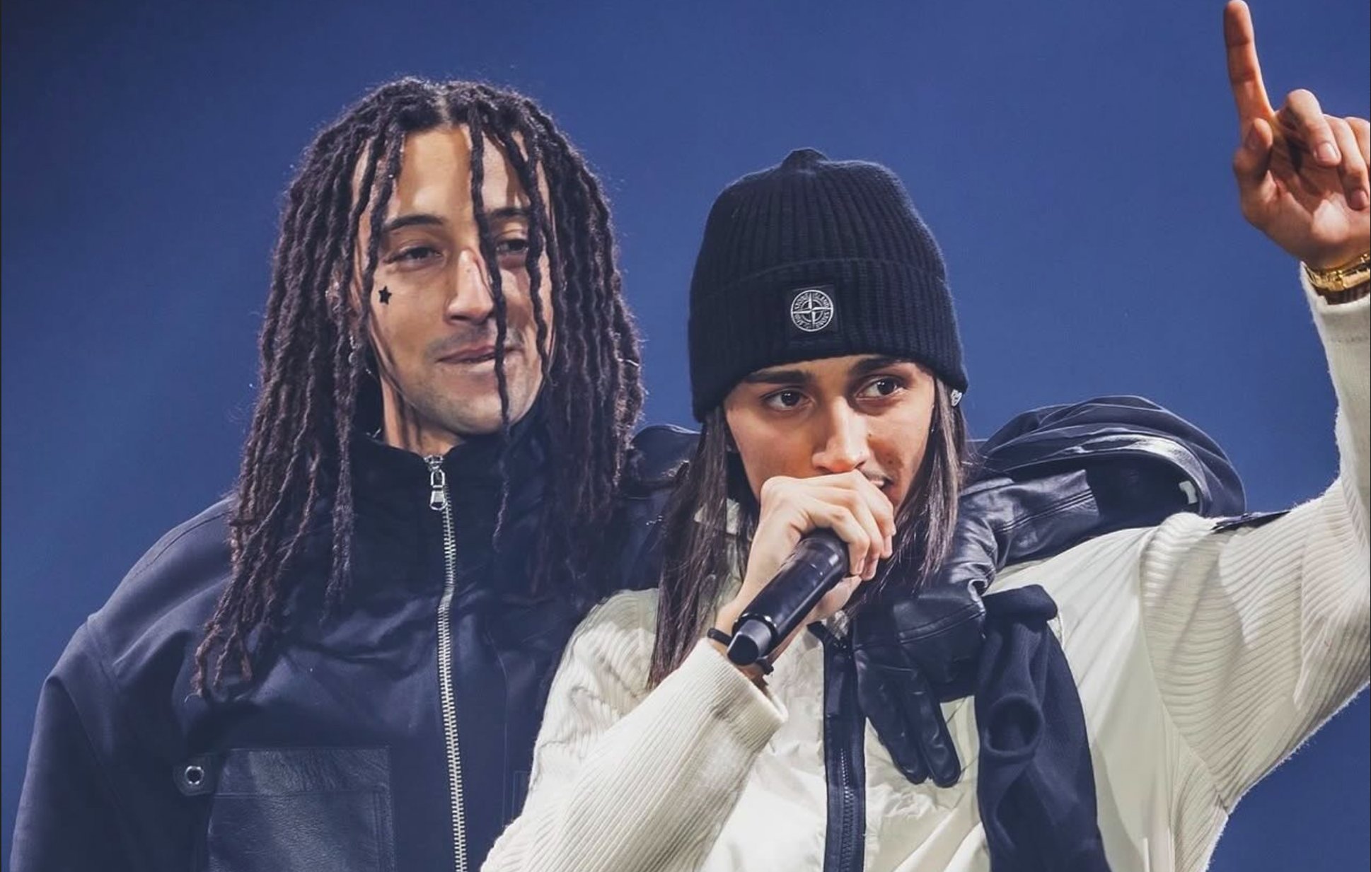 Two performers on stage; one speaks into a microphone while the other stands behind.