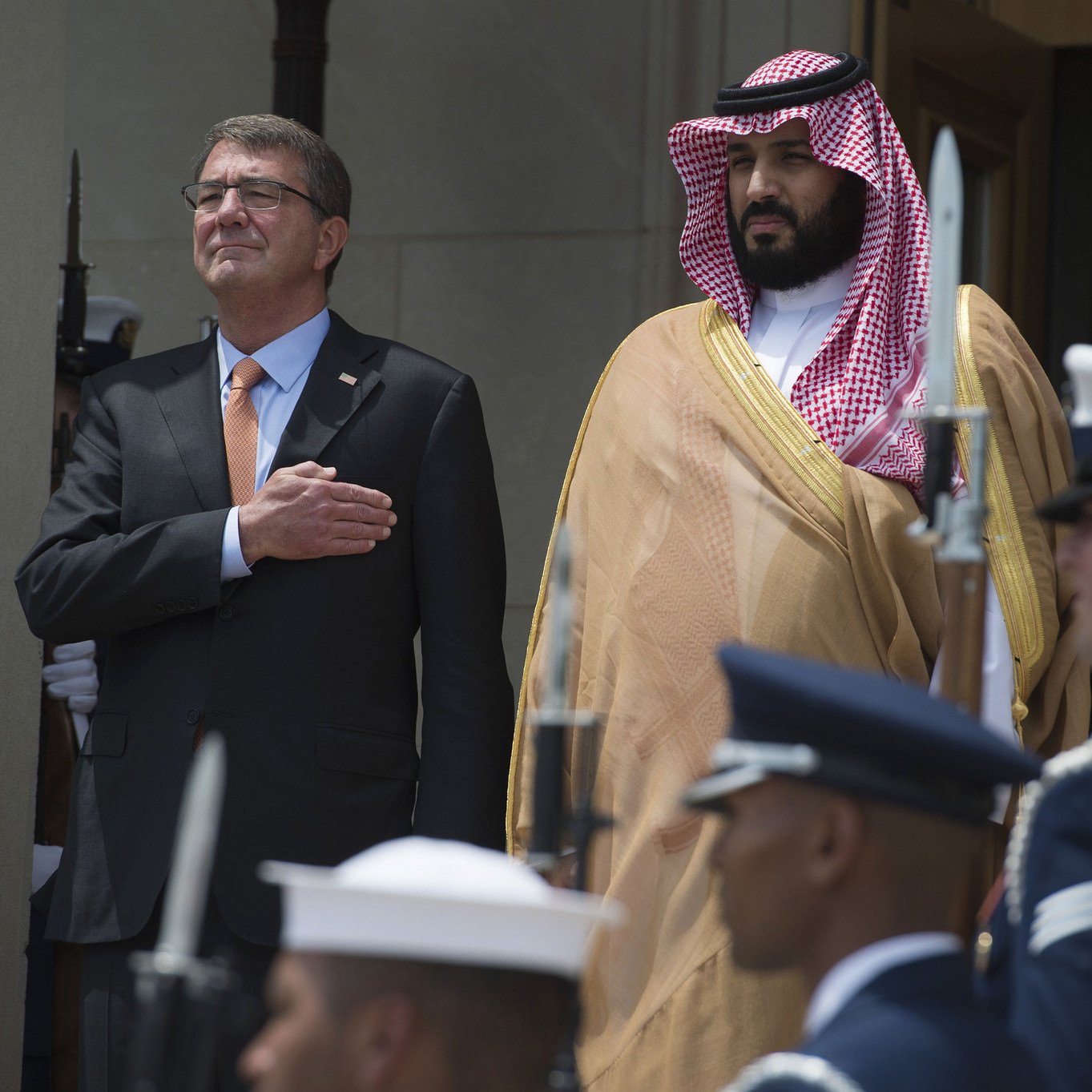 L'image montre deux hommes en tenue formelle. L'un est sur le devant, ayant la main sur le cœur, tandis que l'autre se tient à ses côtés. Ils sont entourés de membres d'une garde d'honneur, portant des uniformes militaires. L'environnement semble officiel, probablement un événement diplomatique ou militaire. Les deux hommes ont l'air sérieux et engagés dans la cérémonie.