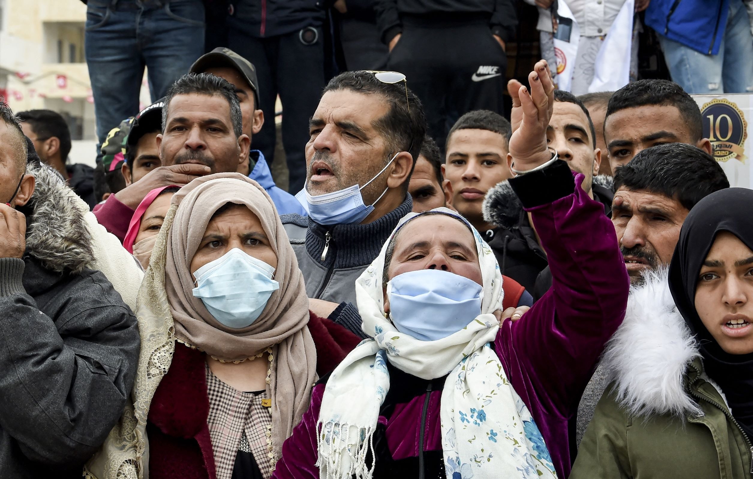 L'image montre une foule de personnes, principalement des hommes et des femmes, réunies pour exprimer leurs émotions. Certaines personnes portent des masques faciaux, et il y a des expressions de détermination et de protestation parmi les membres du groupe. Les vêtements sont variés, avec des accents de couleurs différentes. L'ambiance semble animée et engagée, suggérant une manifestation ou un rassemblement pour une cause.