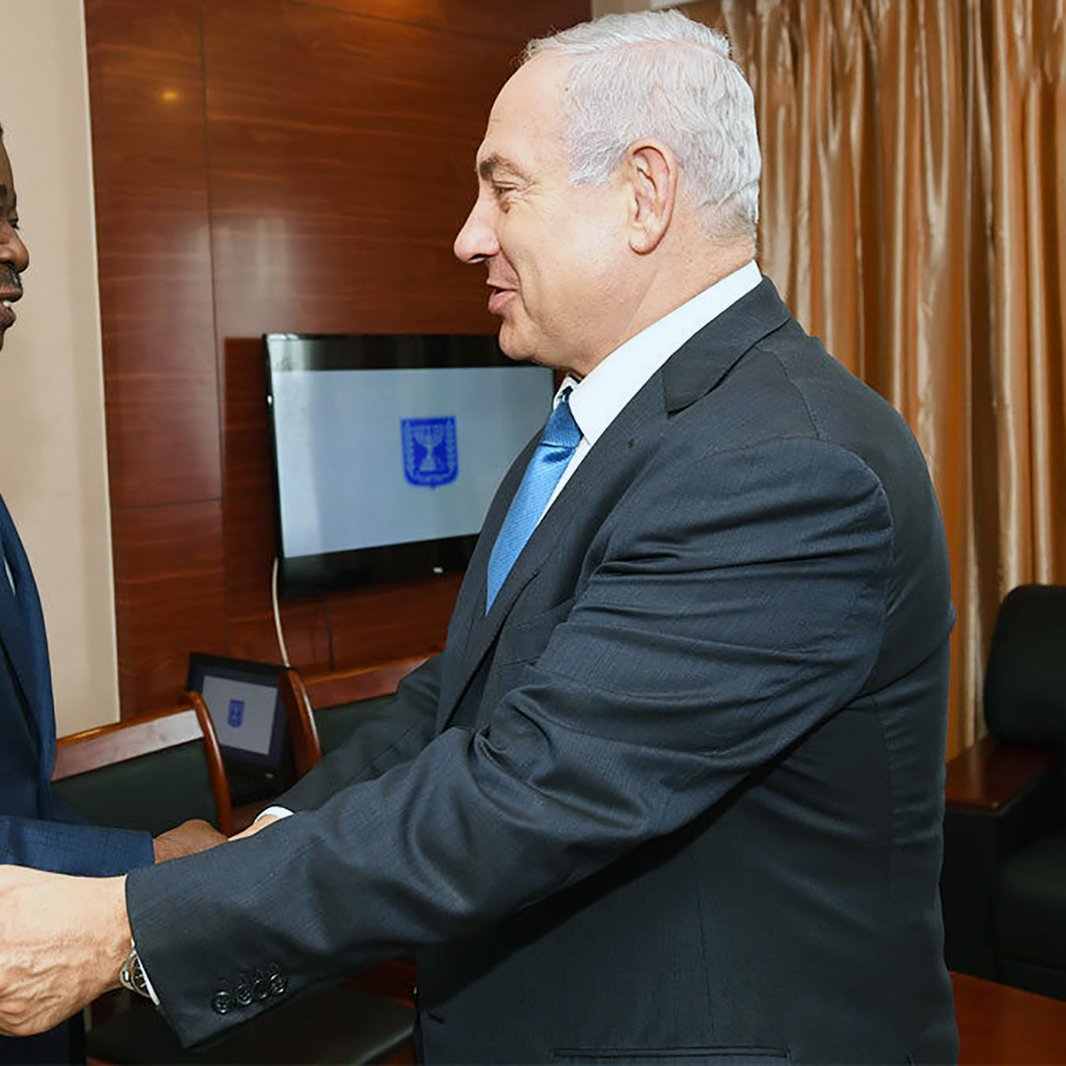 The image depicts two men engaging in a handshake in a formal setting. One man is dressed in a blue suit and tie, while the other is in a dark suit and tie. They appear to be smiling and are facing each other, suggesting a cordial meeting or discussion. The background includes a decorative setting with drapes and some furniture, indicating a professional atmosphere.