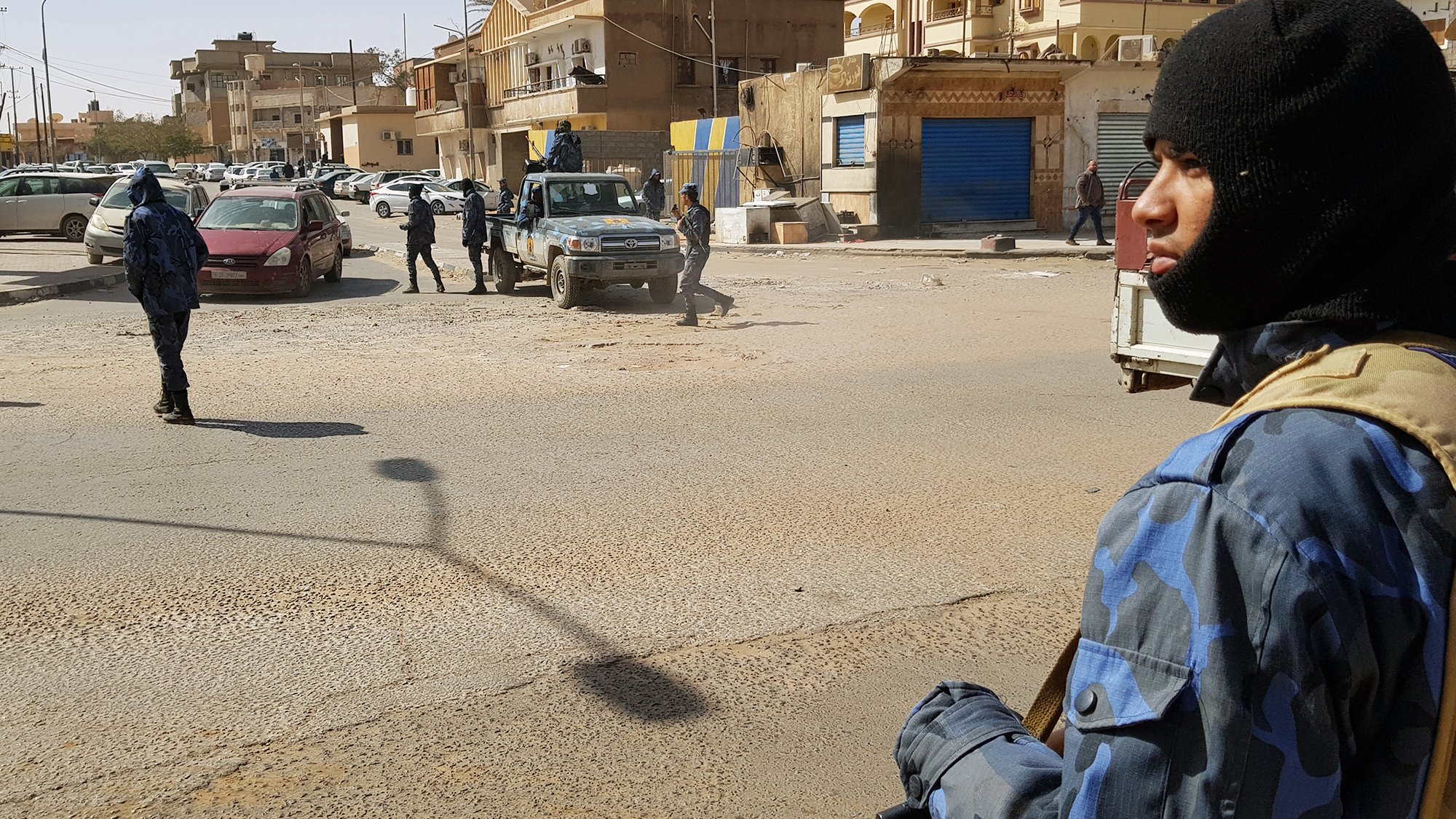 تظهر الصورة مشهداً في منطقة حضرية، حيث يوجد عدد من رجال الأمن يرتدون زيًّا رسميًّا وقبعات سُوداء. أحدهم يحمل سلاحًا ويقف في المقدمة، بينما يتواجد آخرون في الخلفية. تشهد المنطقة حركة مرورية، مع وجود سيارات متوقفة في الشارع. المباني المحيطة تظهر بألوان متنوعة، وبعضها يبدو أنه مغلق. تعكس الصورة أجواءً من اليقظة والحذر الأمني.