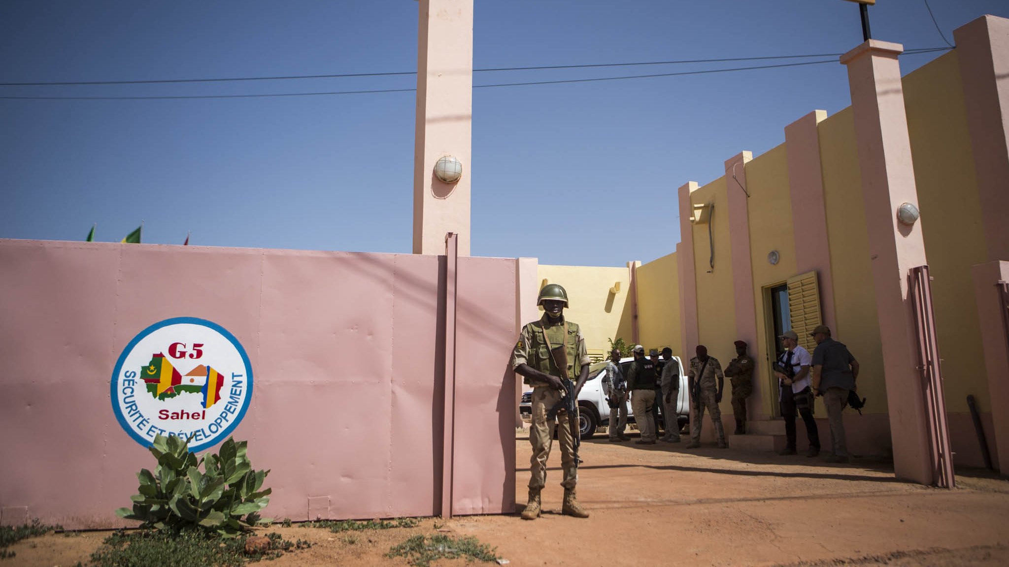 L'image montre un bâtiment qui semble être un poste de commandement de la "Force Conjointe" au sein du G5 Sahel. À l'entrée, on voit un mur avec le logo du G5, ainsi qu'un panneau indiquant "FORCE CONJOINTE POSTE DE COMMANDEMENT". Un soldat en uniforme est en position de garde près de l'entrée, tandis que d'autres personnes sont visibles à l'intérieur ou autour du bâtiment. L'environnement est désertique, et le ciel est dégagé.