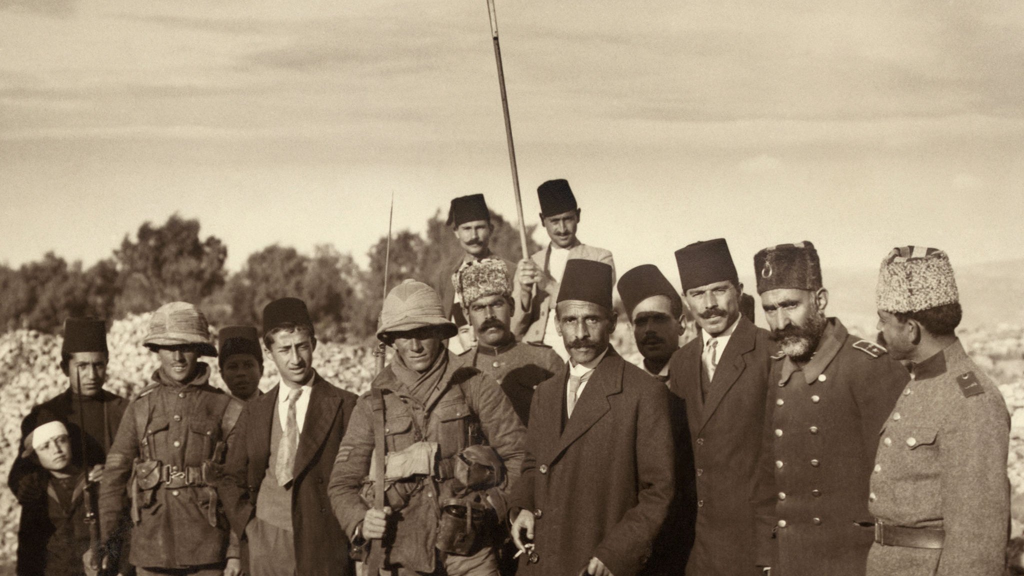 این تصویر تاریخی گروهی از افراد را در یک جایی نشان میدهد. آنها در حال ایستادهاند و برخی از آنها لباسهای نظامی و دیگران لباسهای رسمی و سنتی به تن دارند. یکی از افراد با پرچم سفیدی در مرکز تصویر ایستاده است. محیط اطراف شامل زمینهای ناهموار و درختان است. این تصویر نشاندهنده یک لحظه مهم در تاریخ است و میتواند به یک رویداد خاص یا جمع آوری نیروهای نظامی اشاره داشته باشد.