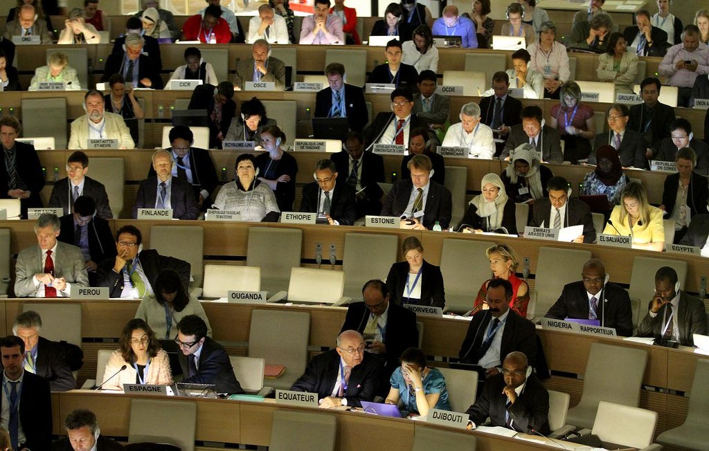 L'image montre une grande salle de conférence remplie de personnes assises à des tables. Les participants semblent concentrés sur leurs ordinateurs portables et divers documents, suggérant qu'ils sont engagés dans une discussion ou une réunion importante. À l'arrière-plan, on peut voir des drapeaux de différents pays visibles, ce qui indique un contexte international. L'éclairage de la salle est tamisé, créant une atmosphère sérieuse et studieuse.