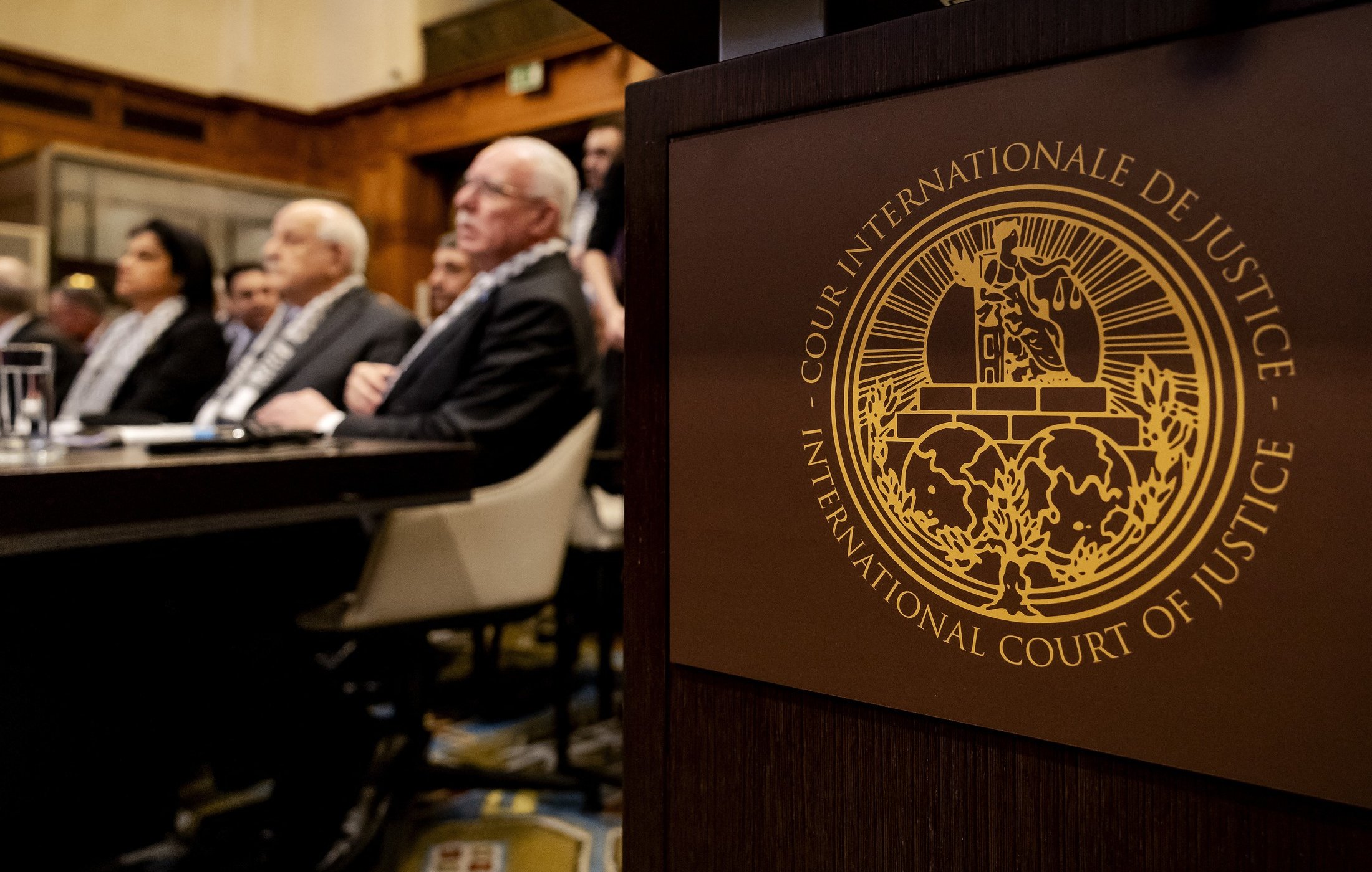 L'immagine mostra un incontro che si svolge in un ambiente formale, probabilmente in un'aula di tribunale. In primo piano si vede un simbolo dell'International Court of Justice (Corte Internazionale di Giustizia), con dettagli dorati che rappresentano l'autorità e la giustizia. Nel background si notano alcune persone sedute, molte delle quali sono visibili di spalle. L'atmosfera è seria e professionale, suggerendo un'importante deliberazione legale.