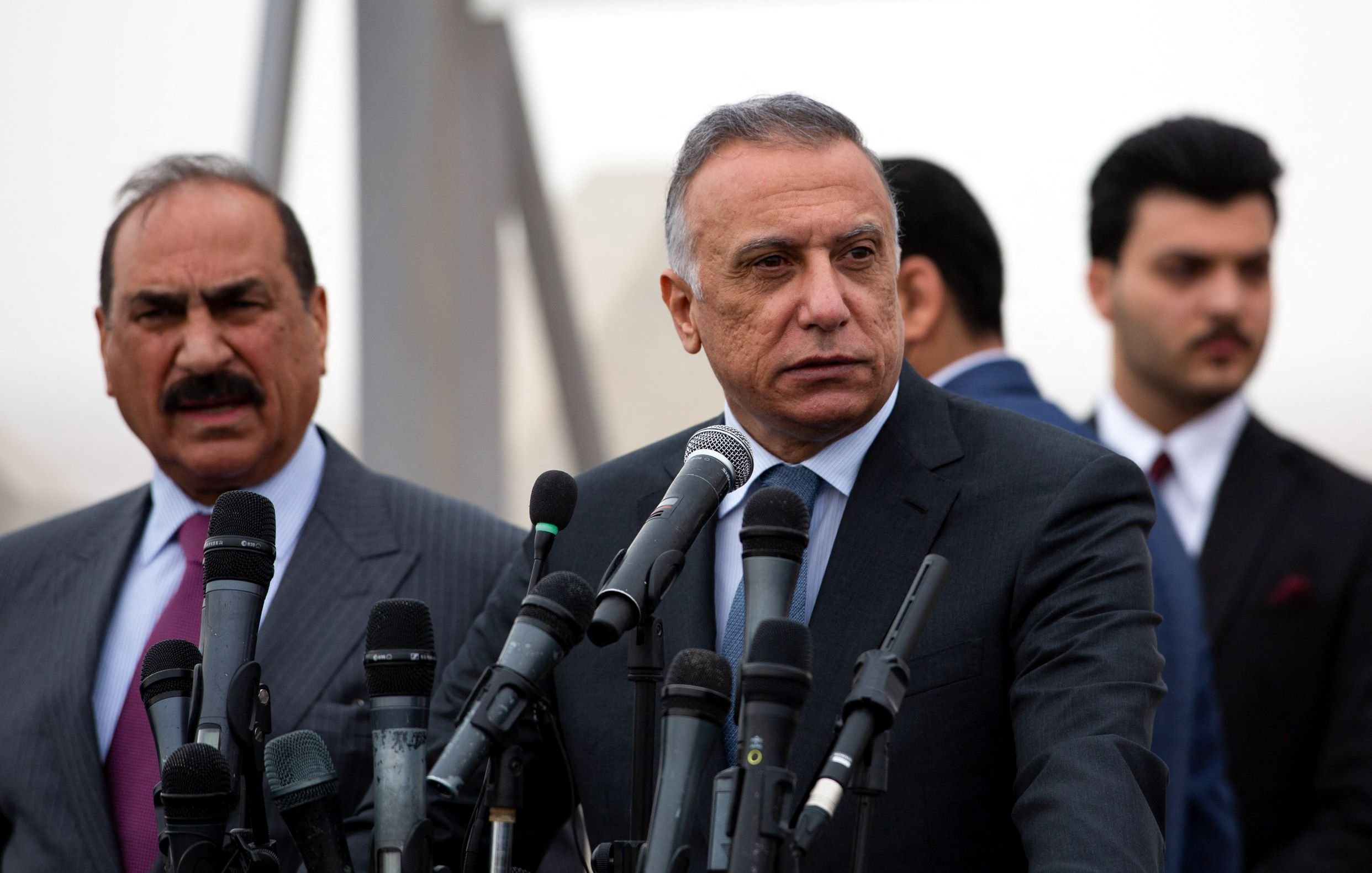 در این تصویر، دو فرد در یک کنفرانس خبری یا مراسم رسمی دیده می‌شوند. فردی که در سمت راست قرار دارد، لباس رسمی پوشیده و به نظر می‌رسد در حال پاسخ دادن به سوالات یا صحبت کردن است. او چند میکروفن مقابل خود دارد که نشان از اهمیت موضوع دارد. فرد دیگر در سمت چپ نیز لباس رسمی دارد و در حال گوش دادن به صحبت‌های فرد اول است. پس‌زمینه تصویر کمی مبهم به نظر می‌رسد. وضوح و جزئیات چهره‌ها به دلیل زاویه و ترکیب تصویر محدود است.