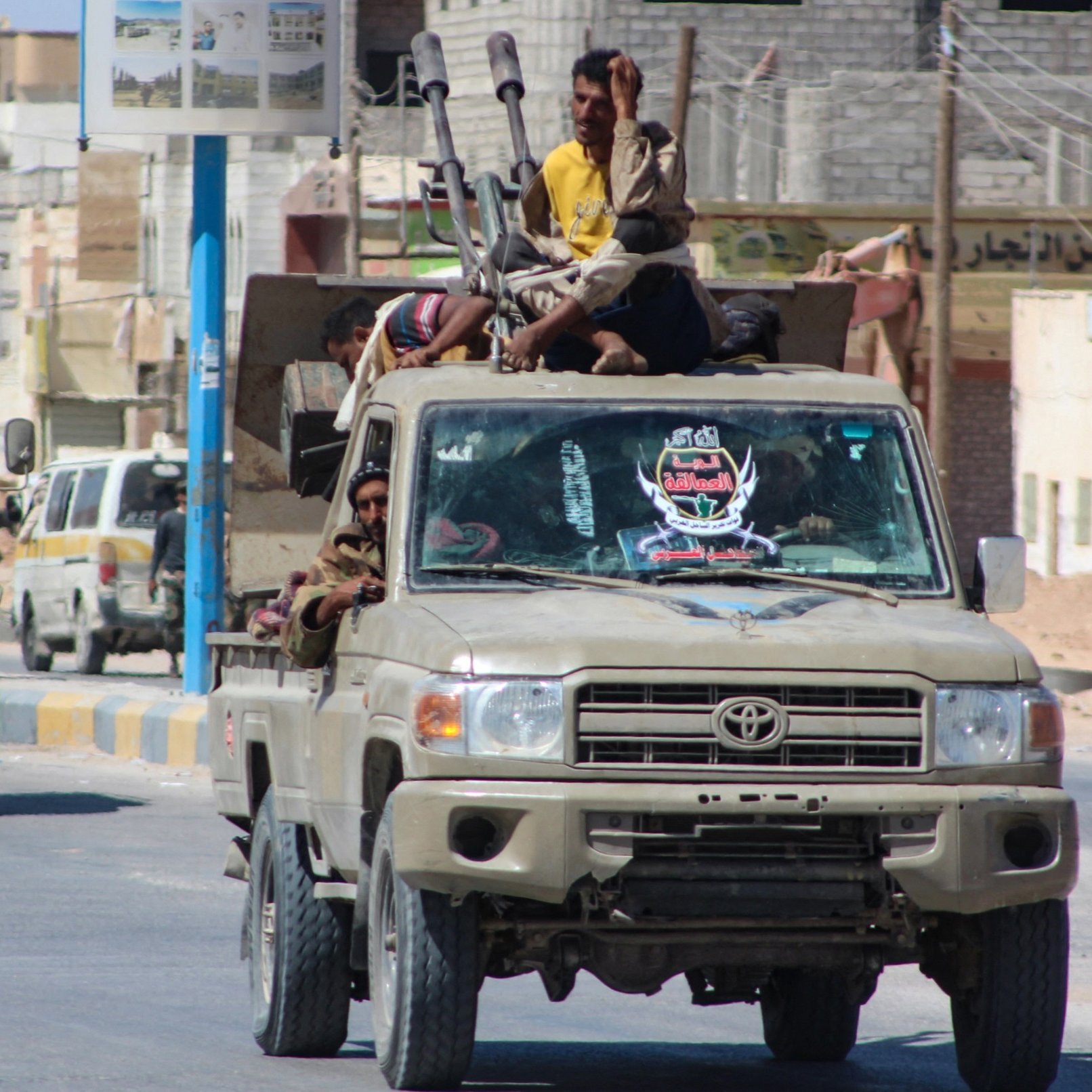 La imagen muestra un convoy de vehículos militares, principalmente camiones, en un ambiente urbano. En uno de los camiones, varias personas están sentadas y de pie en la parte trasera. Algunos están armados y parecen estar en posición de vigilancia. Los vehículos tienen un color beige o khaki, típico de vehículos militares. En el fondo, se pueden ver edificios y una carretera, lo que indica que están en una zona urbana. La escena transmite una sensación de actividad militar en la calle.
