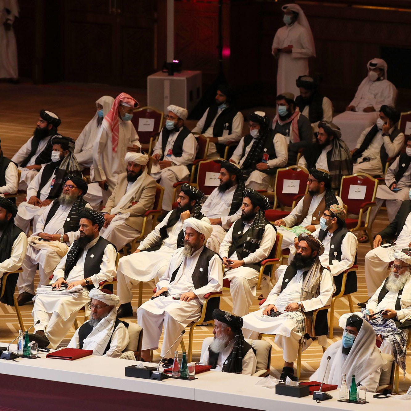 The image depicts a large gathering of individuals seated in a formal setting, likely during a conference or meeting. The attendees are predominantly dressed in traditional attire, including long robes and turbans. Some individuals are wearing face masks, suggesting a concern for health or safety. The arrangement indicates a structured seating plan, with a few individuals at the front possibly in leadership roles. The venue appears to be elegant, with ornate decorations and a spacious layout typical of an official or ceremonial event.