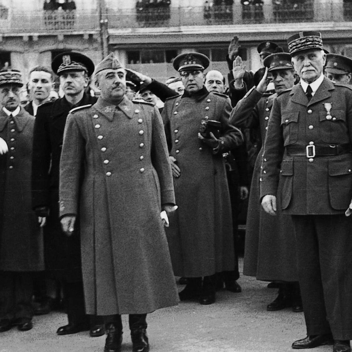 این تصویر سیاه و سفید یک جمعیت نظامی را نشان می‌دهد که شامل تعدادی افسر و مقام‌های عالی‌رتبه است. در مرکز تصویر، دو نفر ایستاده‌اند که به نظر می‌رسد شخصیت‌های مهمی هستند. یکی از آن‌ها در یک کت بلند و با نشان‌های نظامی دیده می‌شود و دیگری نیز لباسی مشابه به تن دارد. افراد دیگر در پس‌زمینه به حالت احترام دست خود را بالا برده‌اند و به نظر می‌رسد در حال ادای احترام هستند. فضای کلی تصویر جدی و رسمی به نظر می‌رسد.