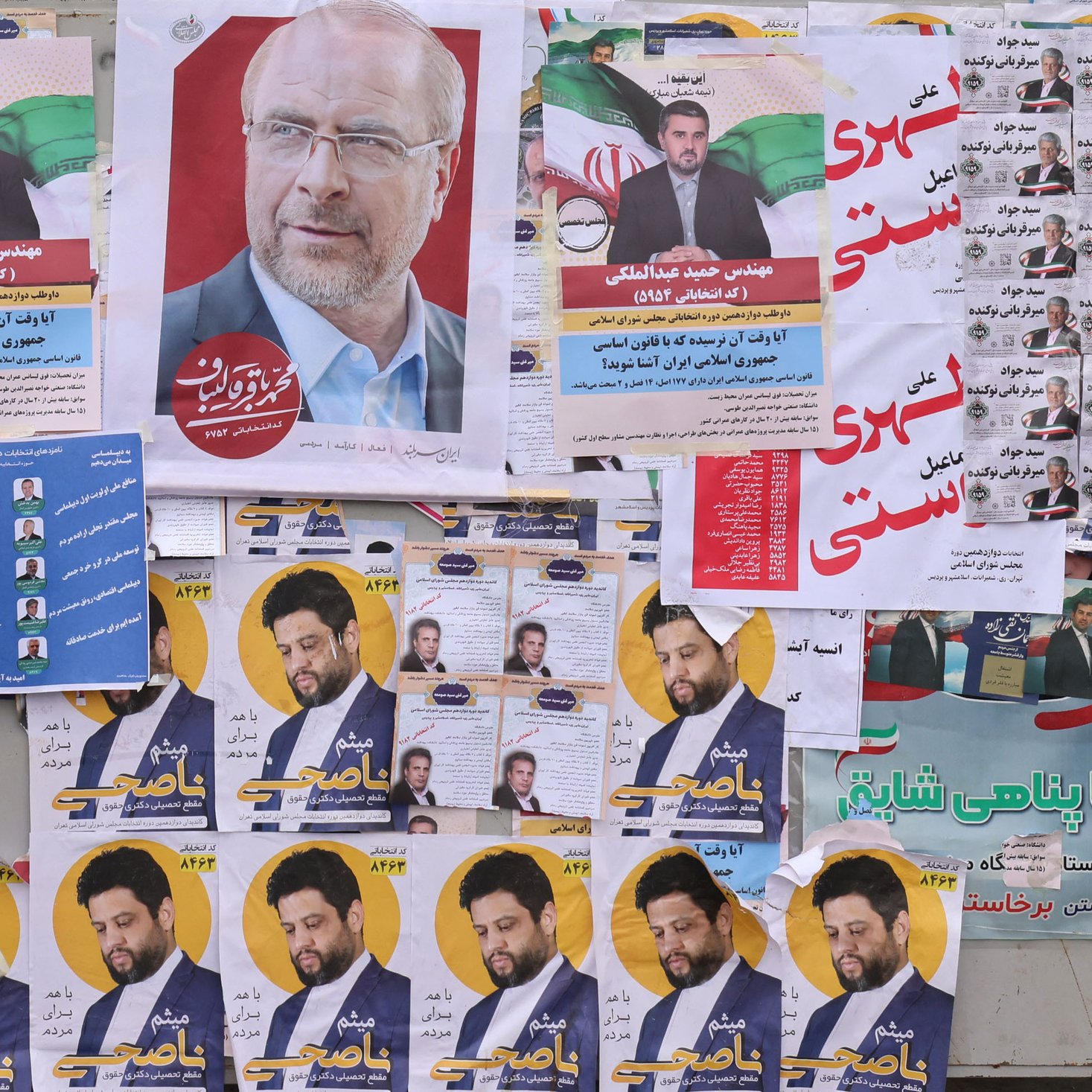 در این تصویر، یک زن که با چادر مشکی پوشیده است، در حال عبور از کنار دیواری است که پر از پوسترهای انتخاباتی و تبلیغاتی است. پوسترها شامل تصاویری از نامزدهای مختلف و شعارهای انتخاباتی هستند. چهره‌ای از یک فرد (احتمالاً نامزد) در مرکز تصویر بسیار مشهود است، در حالی که دیگر پوسترها در اطراف آن به چشم می‌خورند. دیوار پر از رنگ و متن است که به فضای انتخاباتی اشاره می‌کند. زن در حال نگاه کردن به این پوسترها به نظر می‌رسد.