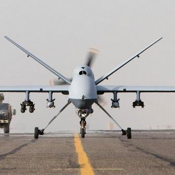 L'image montre un drone militarisé en position de décollage, vu de face, sur une piste d'atterrissage. En arrière-plan, on aperçoit un véhicule militaire qui s'approche. Le ciel semble légèrement nuageux ou brumeux, créant une ambiance de calme avant l'action. Le drone est de couleur claire, avec des ailes larges et un design aérodynamique, prêt à s'envoler.