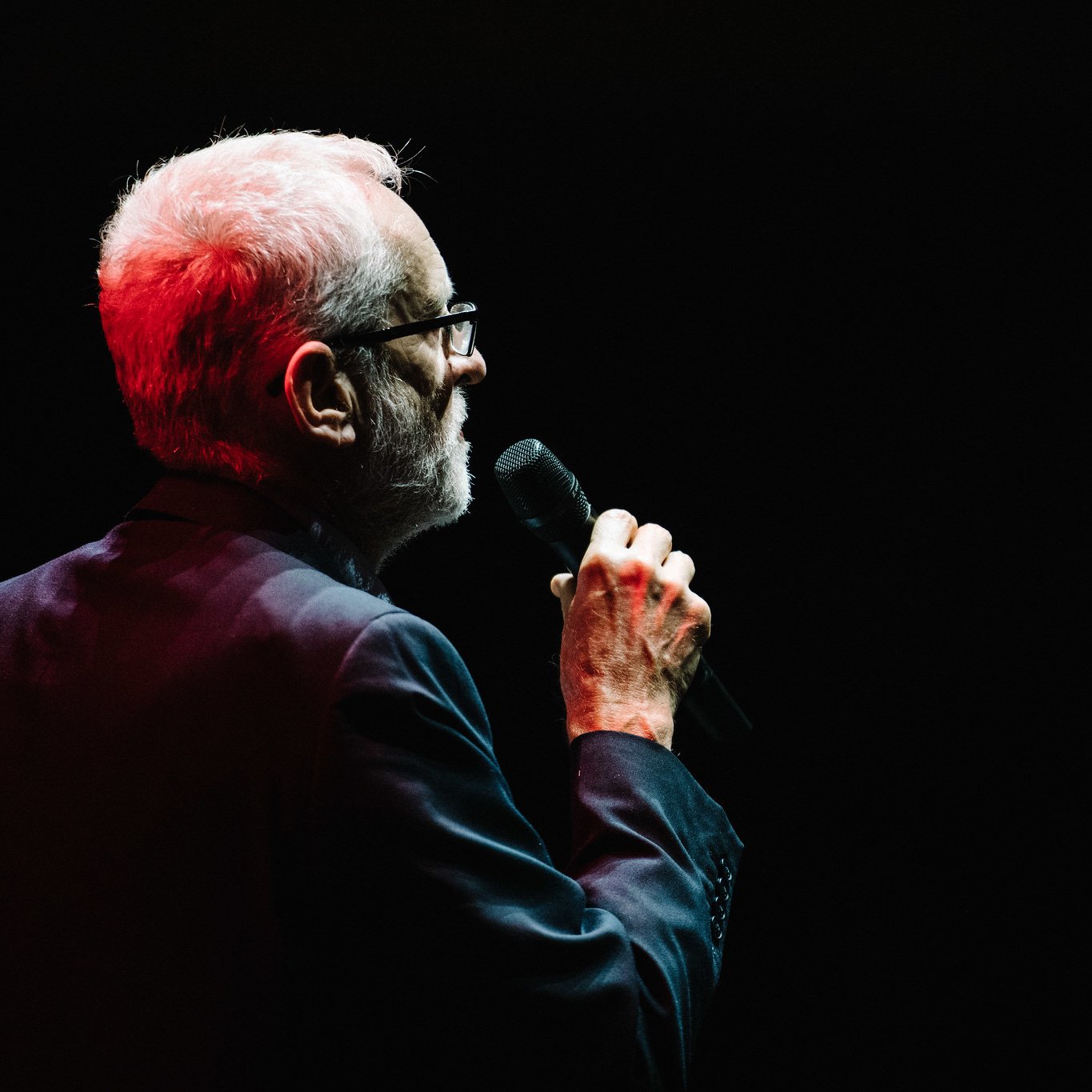 در این تصویر، یک مرد سالخورده دیده می‌شود که از عقب به سمت دوربین ایستاده است. او لباس رسمی و تیره‌ای به تن دارد و عینک زده است. مرد میکروفونی در دست دارد و به نظر می‌رسد در حال سخنرانی یا خواندن است. نور قرمزی از سمت چپ تصویر به او تابیده شده و زمینه تصویر تاریک است که تمرکز را بر روی او بیشتر کرده است.