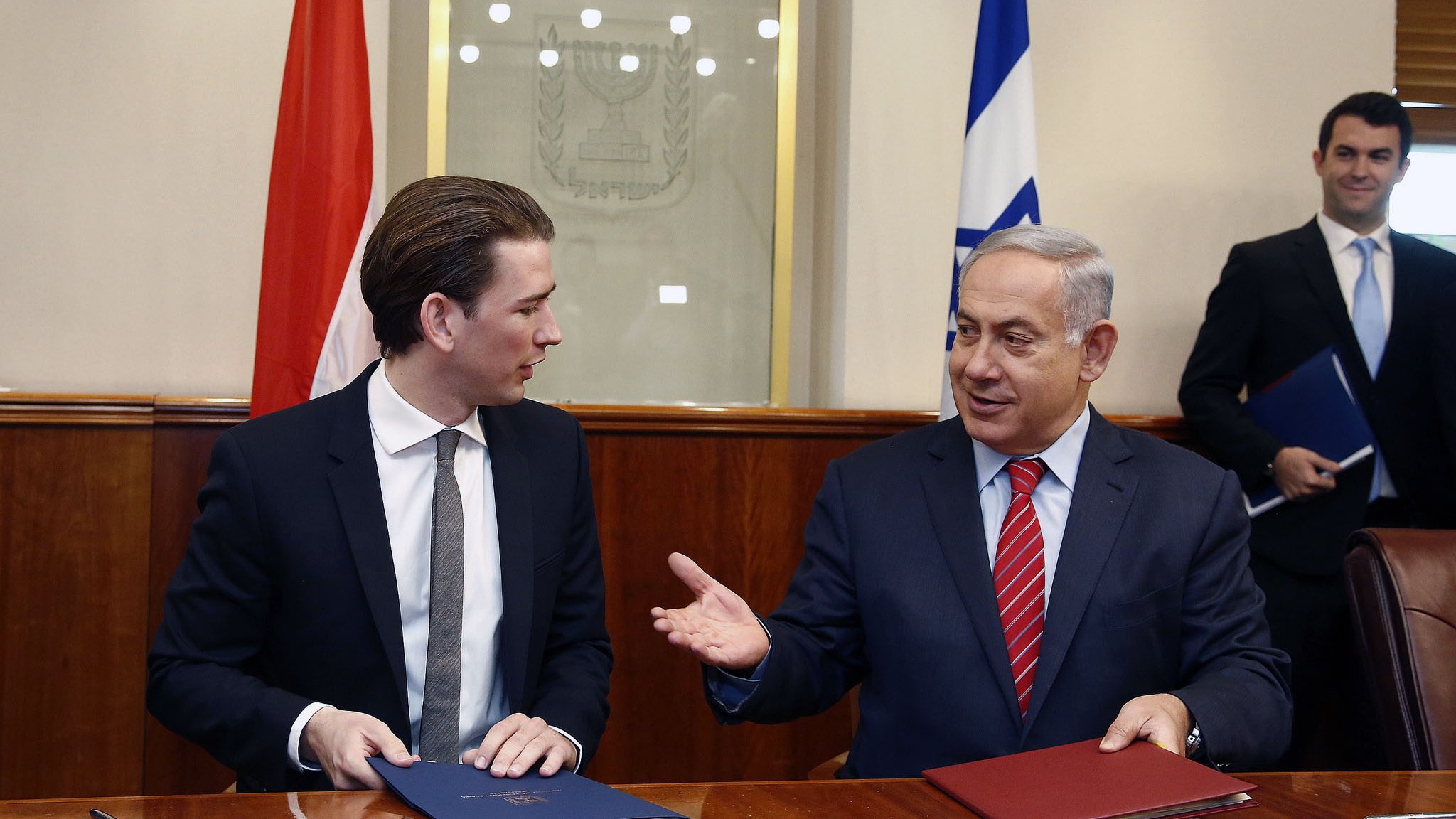 The image shows two men seated at a table in a formal setting. One man, wearing a black suit and tie, appears to be engaged in conversation with the other man, who is wearing a suit with a red tie. They are both holding folders and look to be discussing something important. In the background, there are flags visible, indicating a diplomatic context. The atmosphere appears professional and cooperative.