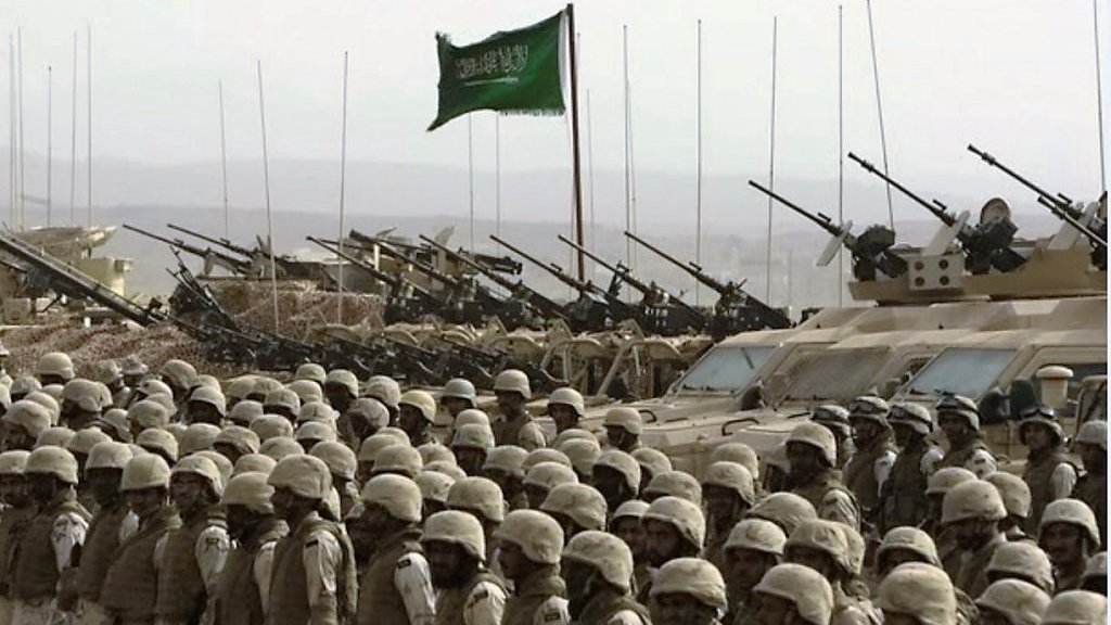 تظهر في الصورة مجموعة من الجنود يرتدون زيًا عسكريًا واقفين في صفوف منتظمة. خلفهم، توجد دبابات ومدرعات عسكرية، حيث تُشاهد المدافع موجهة نحو الأمام. في خلفية الصورة، يوجد علم أخضر يرفرف، مما يوحي بأن هذه الصورة مرتبطة بقوة عسكرية أو استعراض. تعكس الصورة أجواءً عسكرية وهيبة القوات المدافعة.