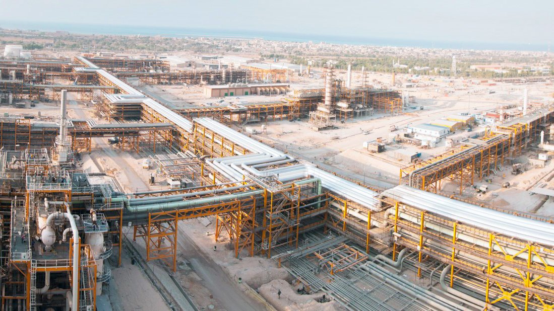 این تصویر یک مجتمع صنعتی بزرگ را نشان می‌دهد که شامل ساختارهای فلزی و لوله‌های گسترده است. در پس‌زمینه، تعدادی سازه بزرگ و تاسیسات به چشم می‌خورد. زمین اطراف به شکل خاکی و خشن است و در دوردست،‌ نوار آبی دریا نمایان است. این مکان به نظر می‌رسد که در حال فعالیت‌های صنعتی مانند پالایش نفت یا گاز باشد. فضا به طور کلی شلوغ و پر از تجهیزات و سازه‌ها است.