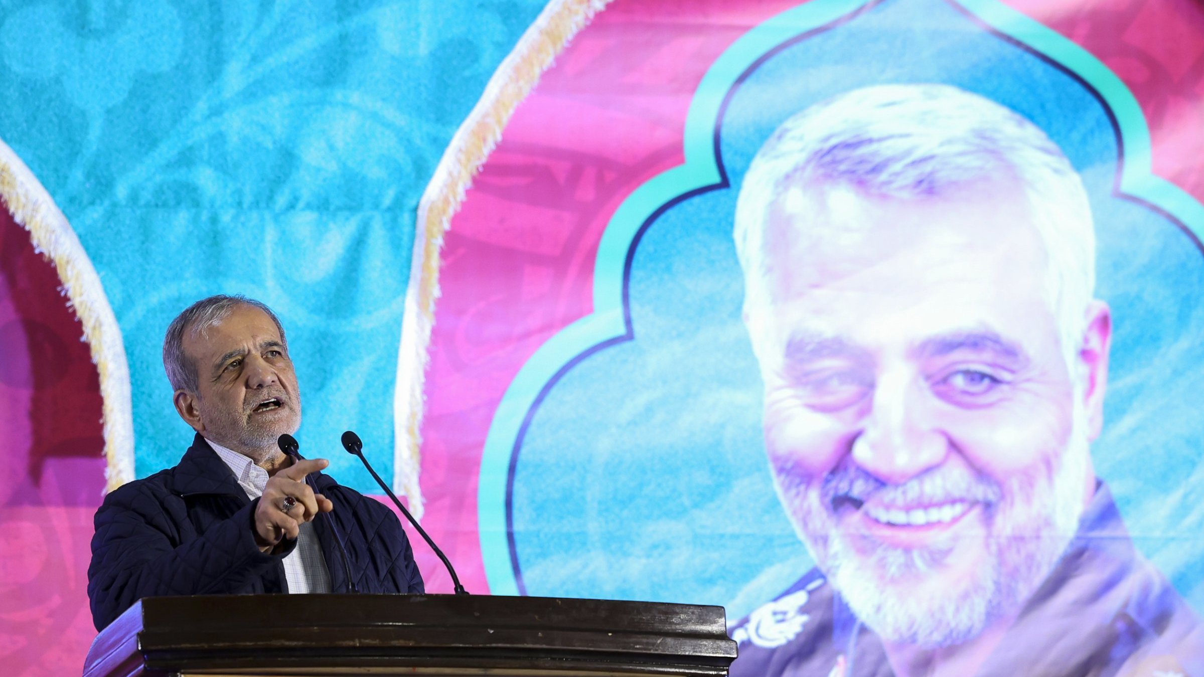 L'image montre un homme en train de s'exprimer lors d'un discours devant un microphone. En arrière-plan, il y a un grand panneau coloré affichant le portrait d'un homme souriant, probablement une figure importante. Le fond est décoré avec des motifs vibrants. L'homme au premier plan semble prendre la parole avec passion, pointant du doigt, ce qui indique qu'il parle probablement d'un sujet important ou émotionnel.