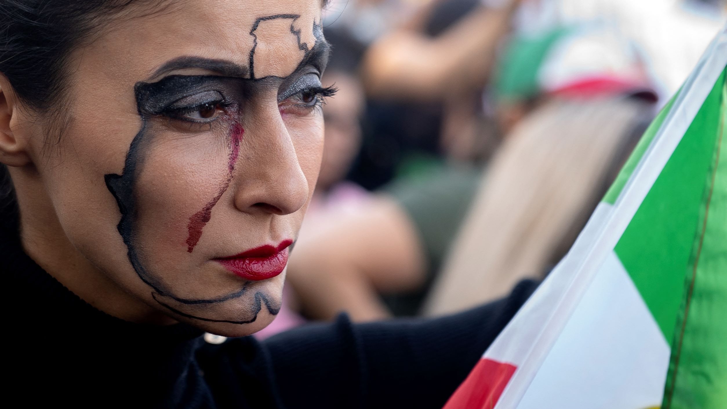 في هذه الصورة، تظهر امرأة تحمل علمًا بألوان الأخضر والأبيض والأحمر. تضع مكياجًا فنيًا على وجهها يتضمن خطوطًا سوداء ودموعًا باللون الأحمر. تعبير وجهها جاد، والمشهد يبدو مليئًا بالناس في الخلفية. الجو العام يشير إلى أن هناك فعاليات أو أحداث تجري في المكان.