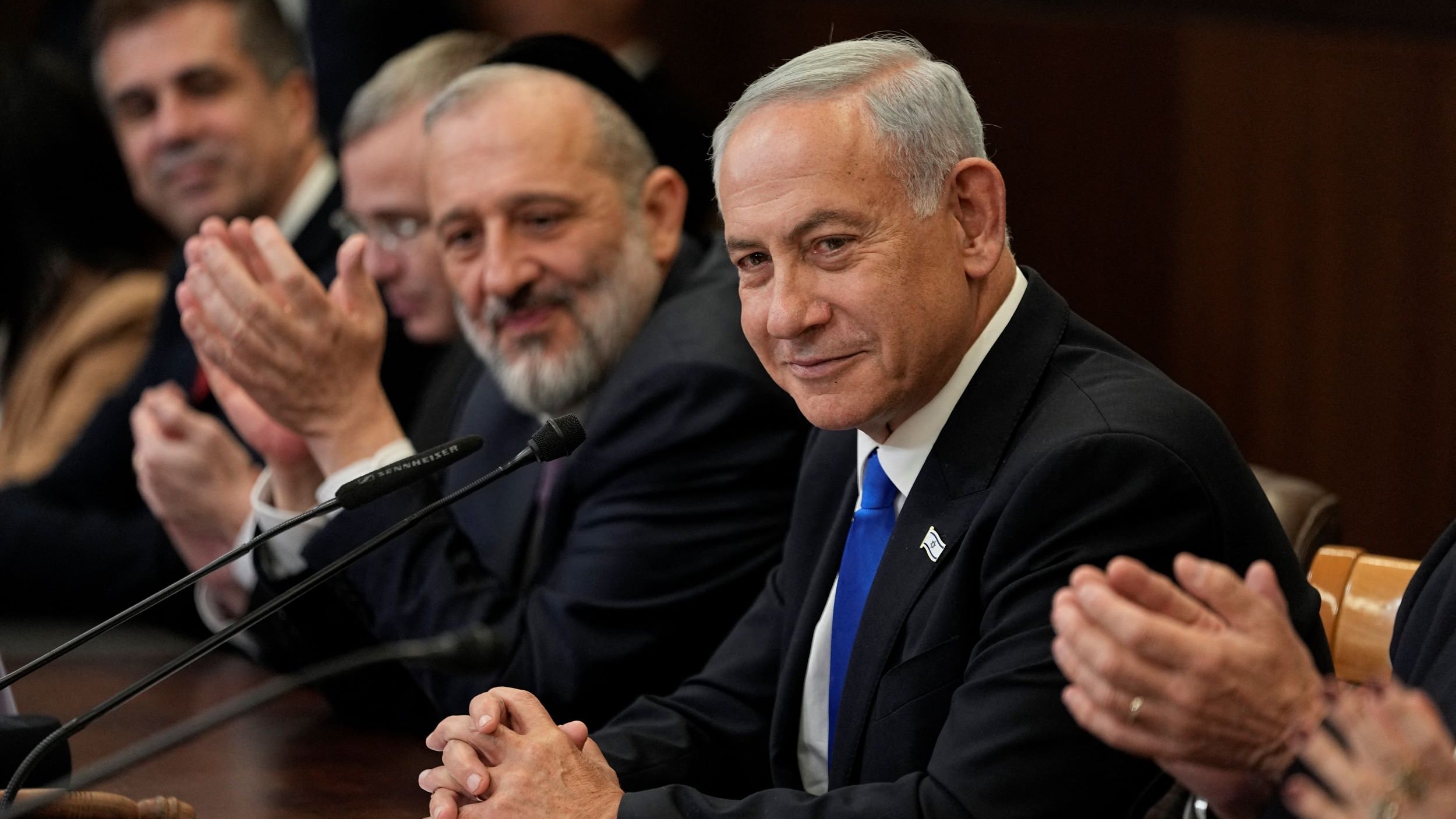 تظهر الصورة مجموعة من الرجال يجلسون حول طاولة اجتماع. في المقدمة، يوجد رجل يرتدي بدلة سوداء وقميص أبيض، يظهر على وجهه تعبير هادئ ويبتسم. إلى جانبه، يجلس رجل آخر يبدو سعيدًا ويصفق، بينما يظهر رجال آخرون في الخلفية يبدون مشغولين بالحديث أو التصفيق. الخلفية تحتوي على ملامح من قاعة اجتماعات.