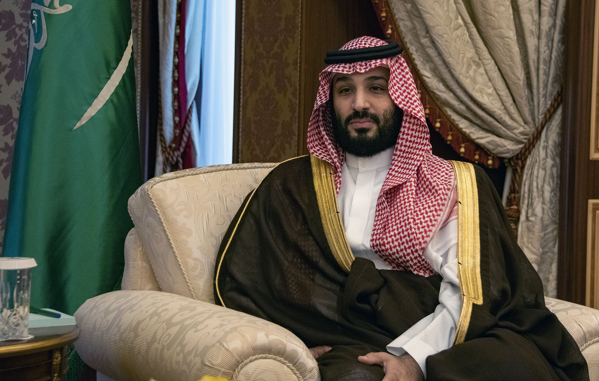L'image montre un homme assis sur un fauteuil, portant une tenue traditionnelle saoudienne, incluant une thobe blanche et un manteau noir avec des détails dorés. Il a une écharpe rouge et blanche, typique de la culture saoudienne, et il est entouré d'un décor riche, incluant des rideaux et des éléments décoratifs. On peut également apercevoir un drapeau saoudien en arrière-plan. L'expression de l'homme semble calme et posée.