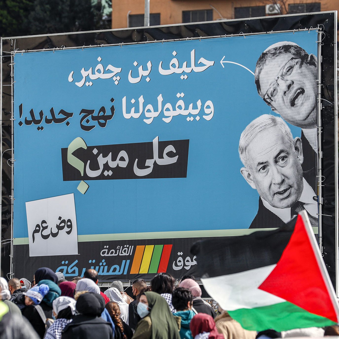 L'image montre une grande affiche sur un panneau publicitaire, présentant des portraits de deux hommes, dont l'un semble être un homme politique conversationnel. En arrière-plan, on peut voir une foule de personnes, certaines tenant des drapeaux palestiniens. L'affiche semble véhiculer un message politique, avec des slogans en arabe, suggérant un contexte de mobilisation ou de protestation. L'ambiance est dynamique, reflétant une situation de rassemblement public.