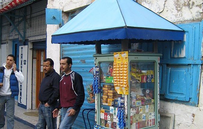 L'image montre un kiosque de vente situé sur un trottoir. Le kiosque, de style ancien, est peint en bleu et abrite divers articles comme des snacks, des boissons et des produits de consommation courante, visibles à travers les vitres. À côté du kiosque, il y a deux hommes. L'un porte un t-shirt, tandis que l'autre est habillé en noir. En arrière-plan, on peut voir des bâtiments aux murs usés et des volets bleus. L'atmosphère a l'air animée et typique d'un marché ou d'une rue commerçante.
