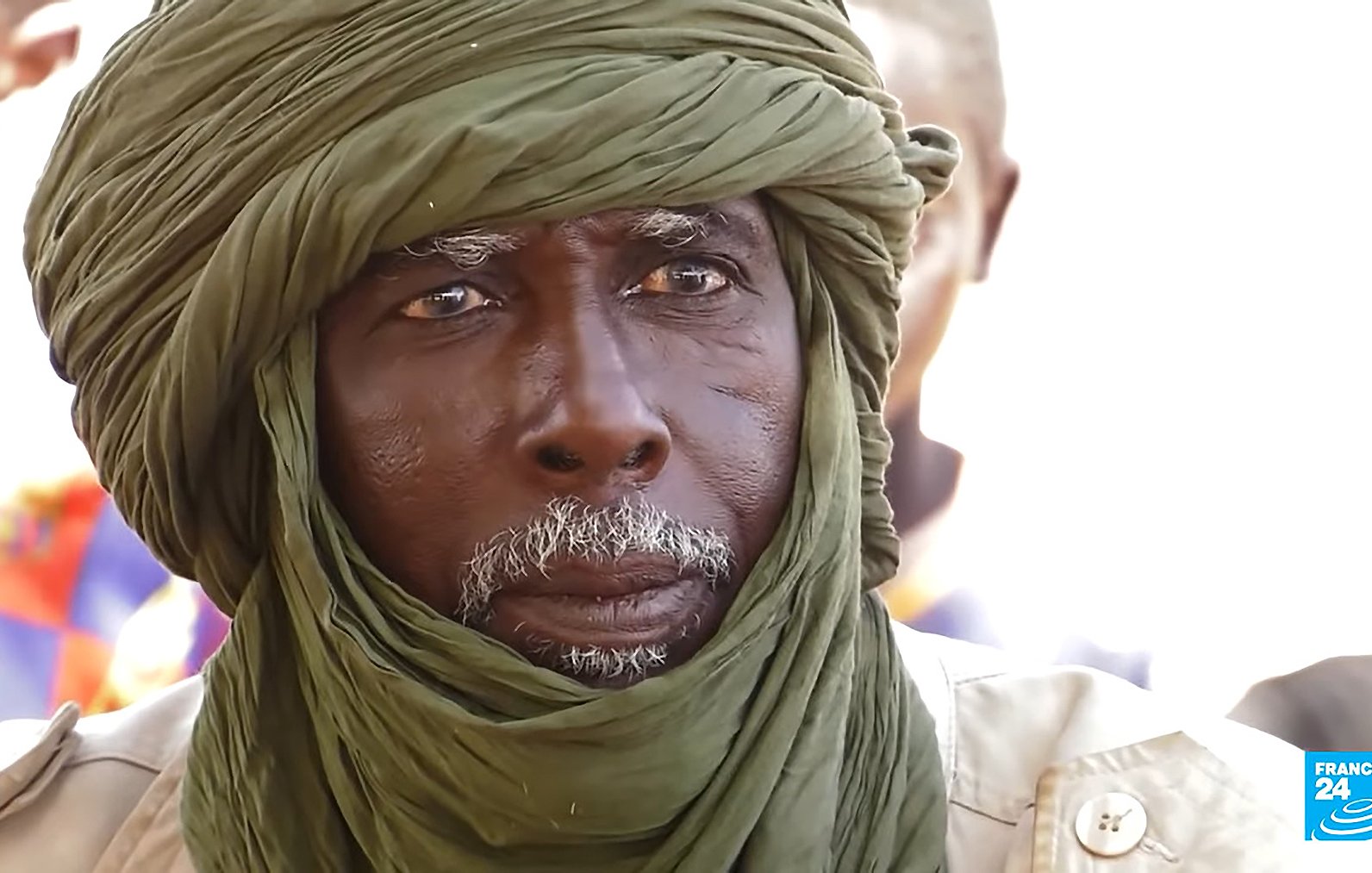The image features an elderly man with a distinguished appearance. He is wearing a green turban wrapped around his head, which adds to his dignified look. His face shows signs of age, with deep wrinkles and a white beard, suggesting wisdom and experience. The background appears to be a social setting with other individuals present, though they are not clearly defined in the image. The focus is on the man, capturing a solemn expression that hints at deep thought or contemplation.