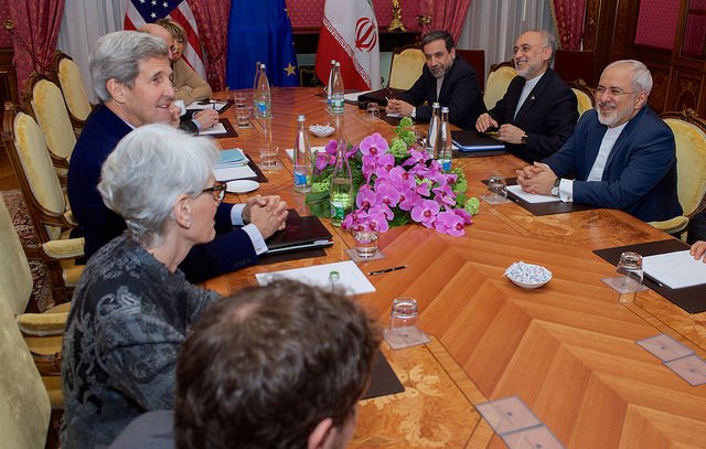 L'image montre une réunion diplomatique autour d'une grande table en bois. Des personnes sont assises de chaque côté, impliquées dans une discussion. On aperçoit des drapeaux d'États-Unis, de l'Union européenne et d'Iran sur la table. Au centre, il y a un bouquet de fleurs, probablement des orchidées. L'ambiance semble sérieuse mais productive, avec des participants au visage souriant, indiquant un échange positif.