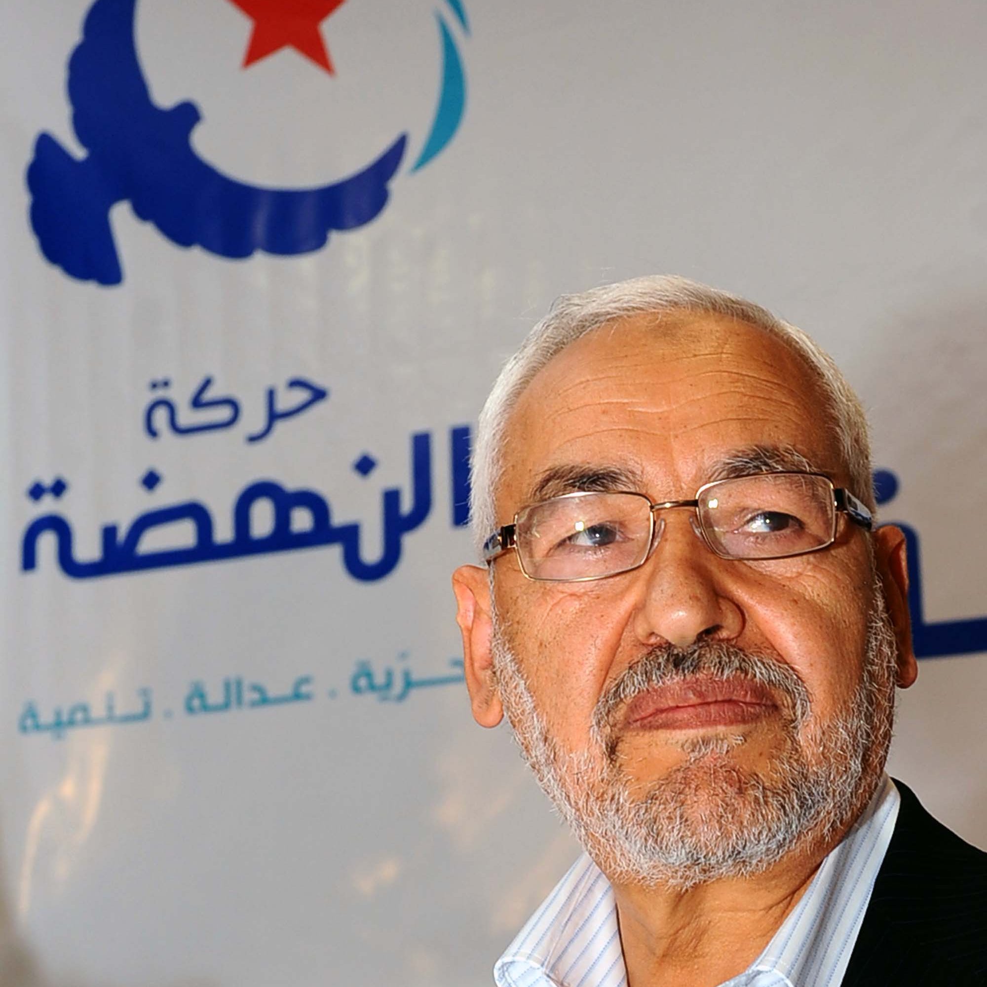 The image features a man with a gray beard and glasses, looking thoughtfully to the side. He is dressed in a light-colored shirt and blazer. In the background, there is a logo with Arabic text and a star, indicating it is related to a political movement or organization. The overall tone of the image suggests a serious or contemplative mood.