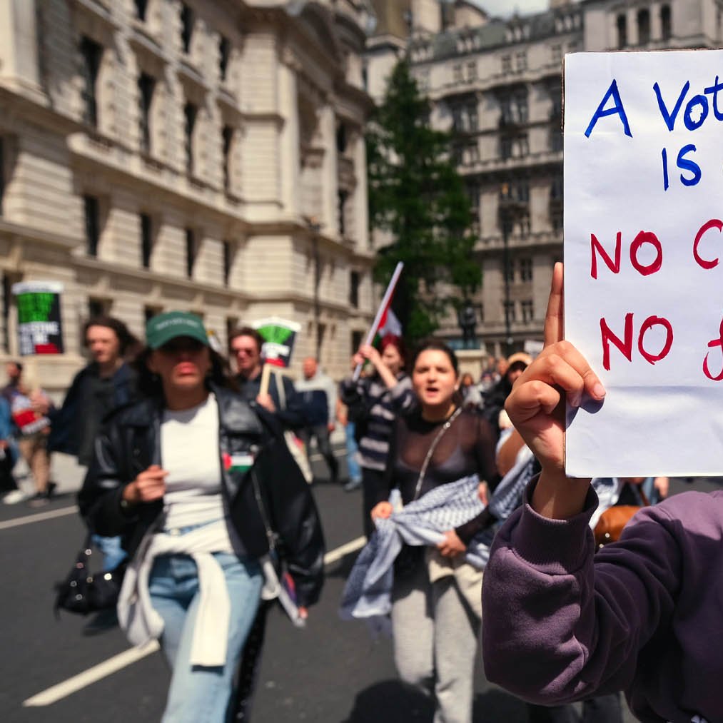 تصویر مربوط به یک تظاهرات است. در جلوی تصویر فردی دیده می‌شود که تابلویی را در دست دارد. روی تابلو نوشته شده: "رای به کارگر، رای به نسل‌کشی است" و همچنین جمله‌ای دیگر به این مضمون که "نه آتش‌بس، نه رای لعنتی!" در پس‌زمینه، جمعیتی از معترضان در حال راهپیمایی دیده می‌شوند. فضای عمومی و شهری است و مردم با شور و هیجان در حال اظهار نظر در مورد موضوعات اجتماعی و سیاسی هستند.