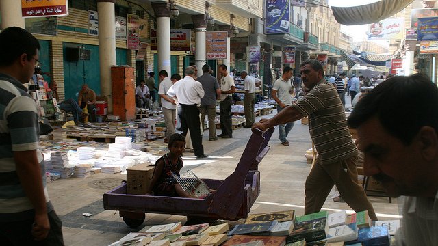 L'image montre un marché animé, probablement un souk, avec des étals de livres disposés au sol. On peut voir plusieurs personnes se déplacer dans la rue, certains feuilletant des livres. Un homme pousse un chariot, où un enfant est assis, observant autour de lui. Les bâtiments en arrière-plan suggèrent une architecture locale, et l'ambiance paraît chaleureuse et vivante.