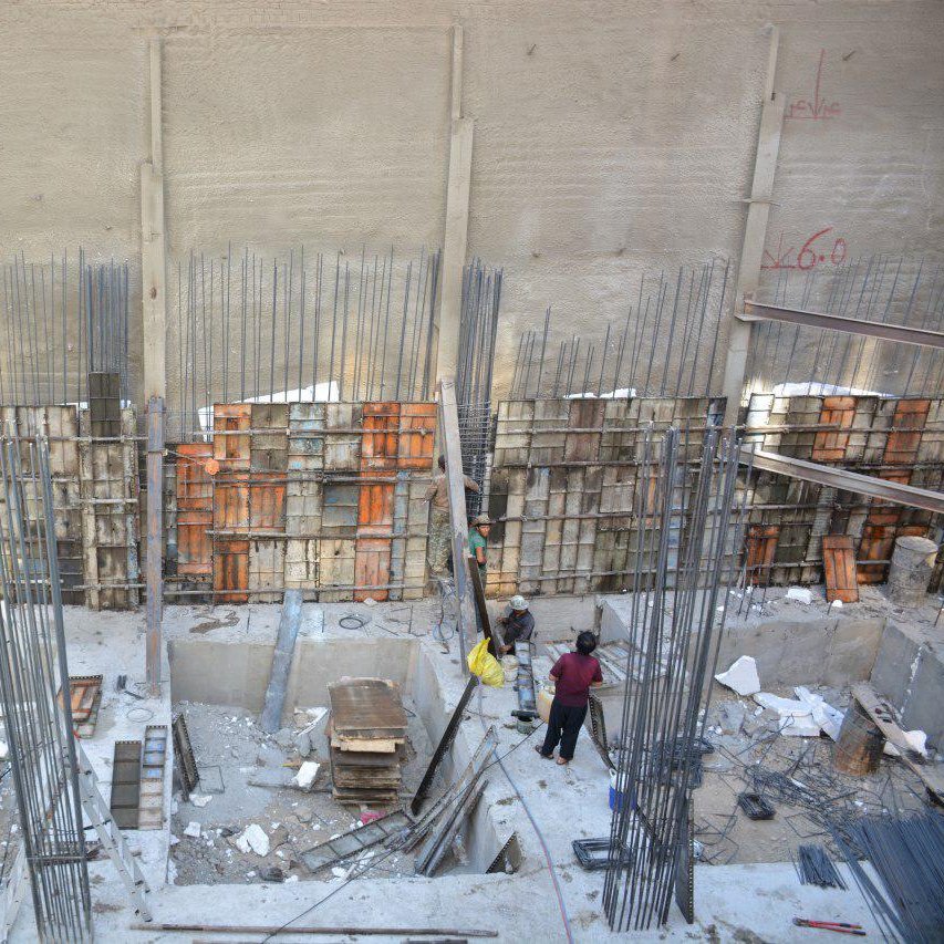 تصویر یک محل construction (ساخت و ساز) را نشان می‌دهد. در این تصویر، یک سازه بتونی در حال انجام است که دیوارهای آن با میله‌های فولادی و قالب‌های چوبی محصور شده‌اند. عده‌ای از کارگران در حال فعالیت و جابجایی مصالح هستند. زمین پروژه نیز برخی مصالح و تجهیزات ساختمانی مانند تخته، میله‌های فولادی و ابزارهای دیگر را نشان می‌دهد. دیوار پشتی با یک سطح صاف و عاری از هرگونه تزئینات، نمایان است.