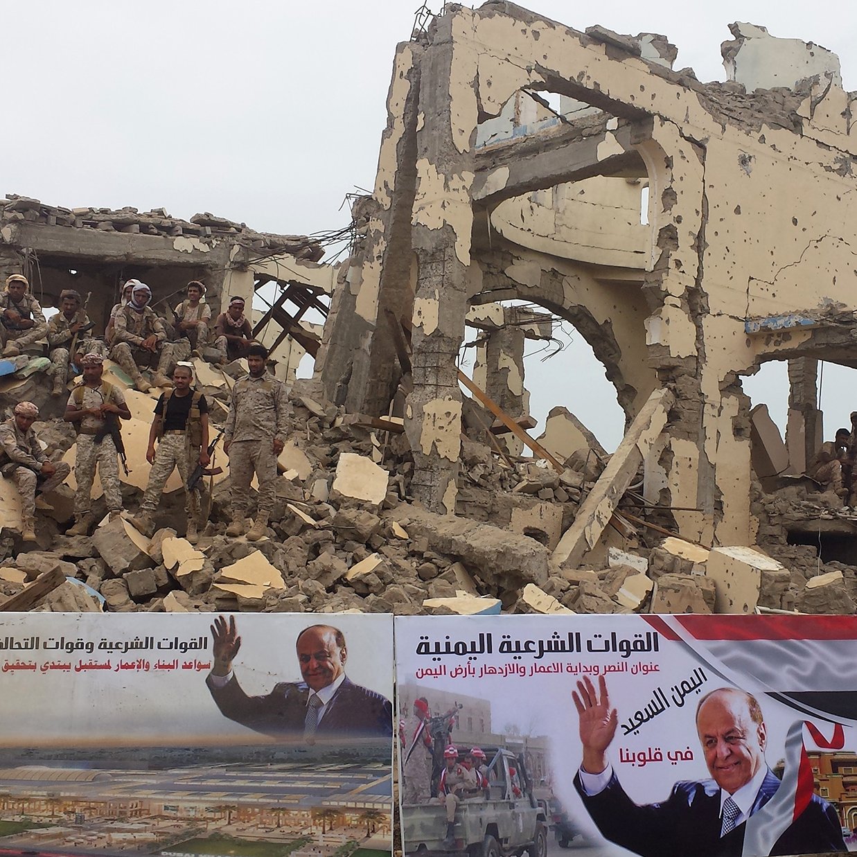 این تصویر نمایی از یک ساختمان ویران‌شده را نشان می‌دهد که تخریب آن به وضوح قابل مشاهده است. در پس‌زمینه، تعدادی نظامی به چشم می‌خورند و روی دیوارهای ویرانه بنرهایی وجود دارد که به نظر می‌رسد تبلیغاتی سیاسی هستند. در این بنرها، تصاویری از پروژه‌ها و شخصیت‌های سیاسی دیده می‌شود. به طور کلی، تصویر به احساساتی از جنگ، تخریب و همچنین تبلیغات سیاسی اشاره می‌کند.