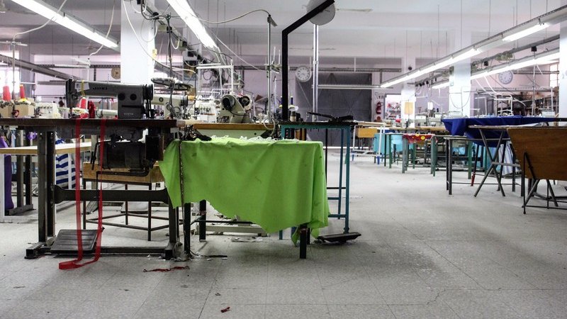 L'image montre un atelier de couture ou de fabrication. On y voit plusieurs tables de travail avec des machines à coudre. Certaines machines semblent être à l'arrêt, et des morceaux de tissu sont éparpillés sur le sol. L'éclairage est assez lumineux, et l'espace semble vaste mais légèrement en désordre, ce qui donne une impression d'activité récente. Des outils et du matériel de couture sont visibles sur les surfaces de travail, et le décor est fonctionnel et utilitaire.