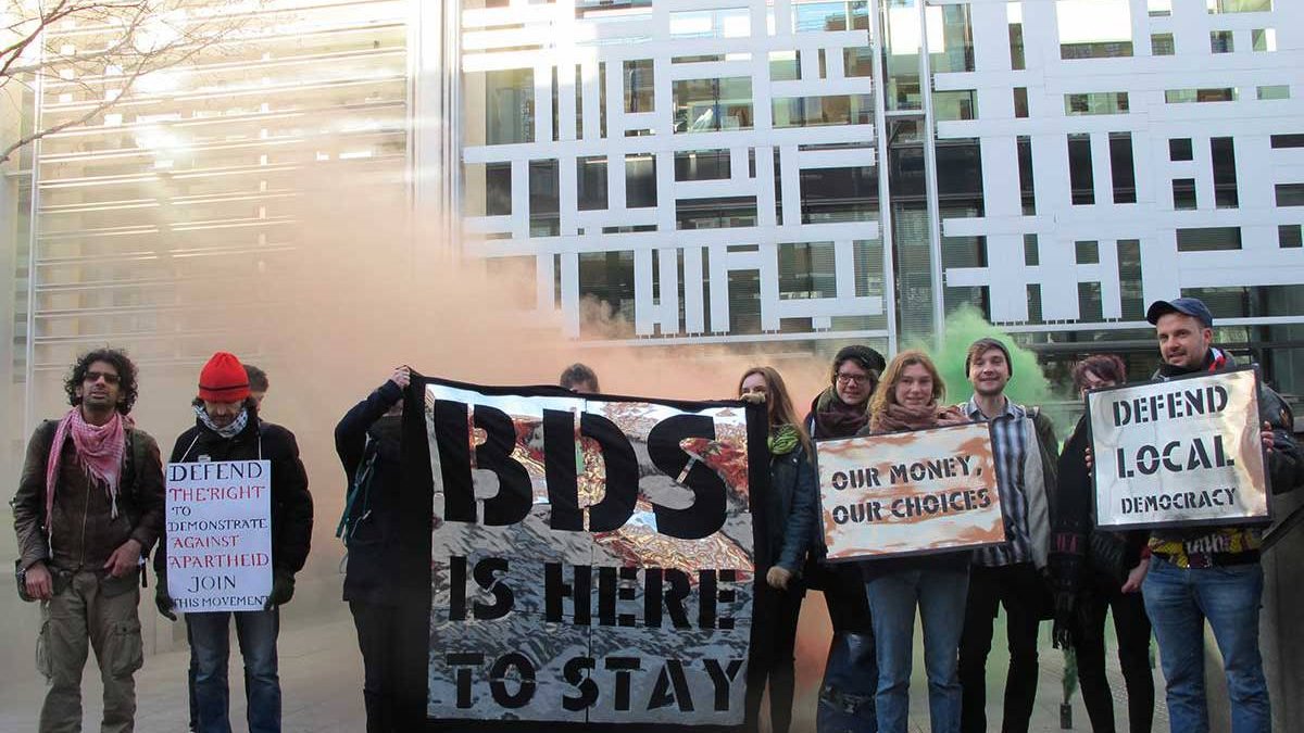 L'image montre un groupe de personnes mobilisées lors d'une manifestation. Elles tiennent différentes pancartes avec des messages engagés, notamment en soutien au mouvement BDS (Boycott, Désinvestissement, Sanctions). L'ambiance semble dynamique et militante, avec des couleurs vives et une légère fumée en arrière-plan, ce qui suggère un moment de tension ou d'énergie. Les participants, vêtus de manière variée, affichent des expressions déterminées face à la caméra.