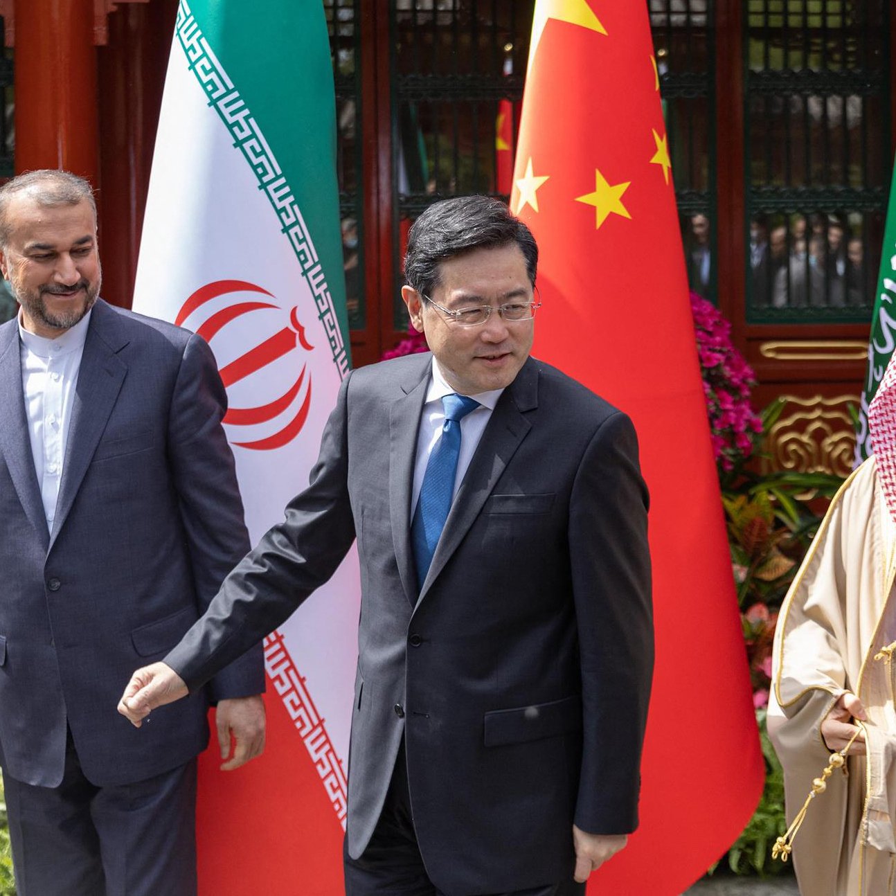 La imagen muestra a tres hombres en un ambiente formal, posiblemente en una reunión diplomática. A la izquierda, un hombre vestido con un traje oscuro parece estar sonriendo ligeramente. En el centro, otro hombre con una corbata azul parece estar en movimiento, mientras que a la derecha, un tercer hombre vestido con una túnica clara y una capa está sonriendo abiertamente. Detrás de ellos se pueden ver banderas de Irán, China y Arabia Saudita, lo que sugiere que esta reunión involucra a estos tres países. El entorno parece ser elegante y bien cuidado.