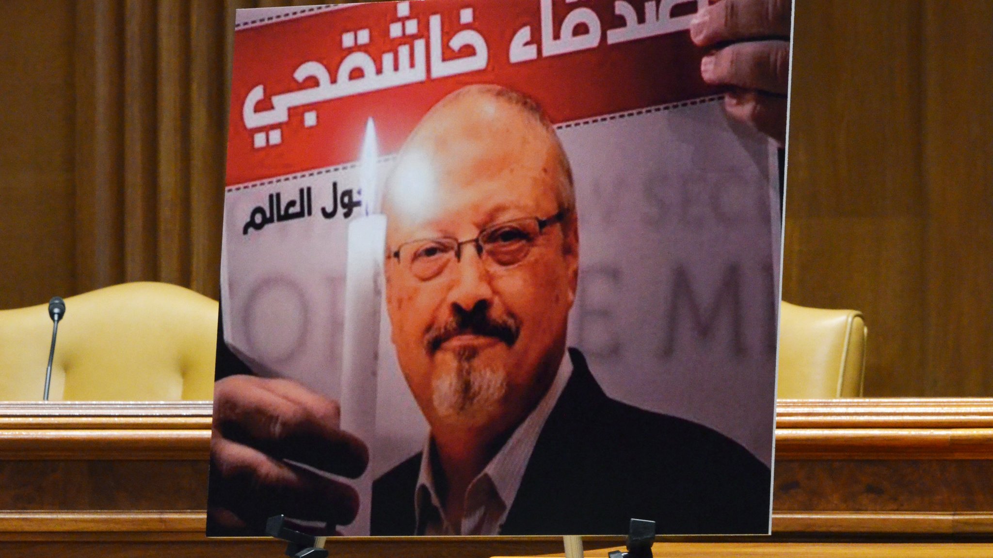 L'image montre un portrait d'un homme tenant une bougie, accompagné d'un texte en arabe et en français. Le fond de l'affiche évoque un hommage ou un soutien, probablement lié à une question de justice. Le contexte global semble être une commémoration ou une manifestation pour défendre les droits humains. Deux chaises sont visibles en arrière-plan, suggérant que l'affiche est présentée lors d'un événement formel.