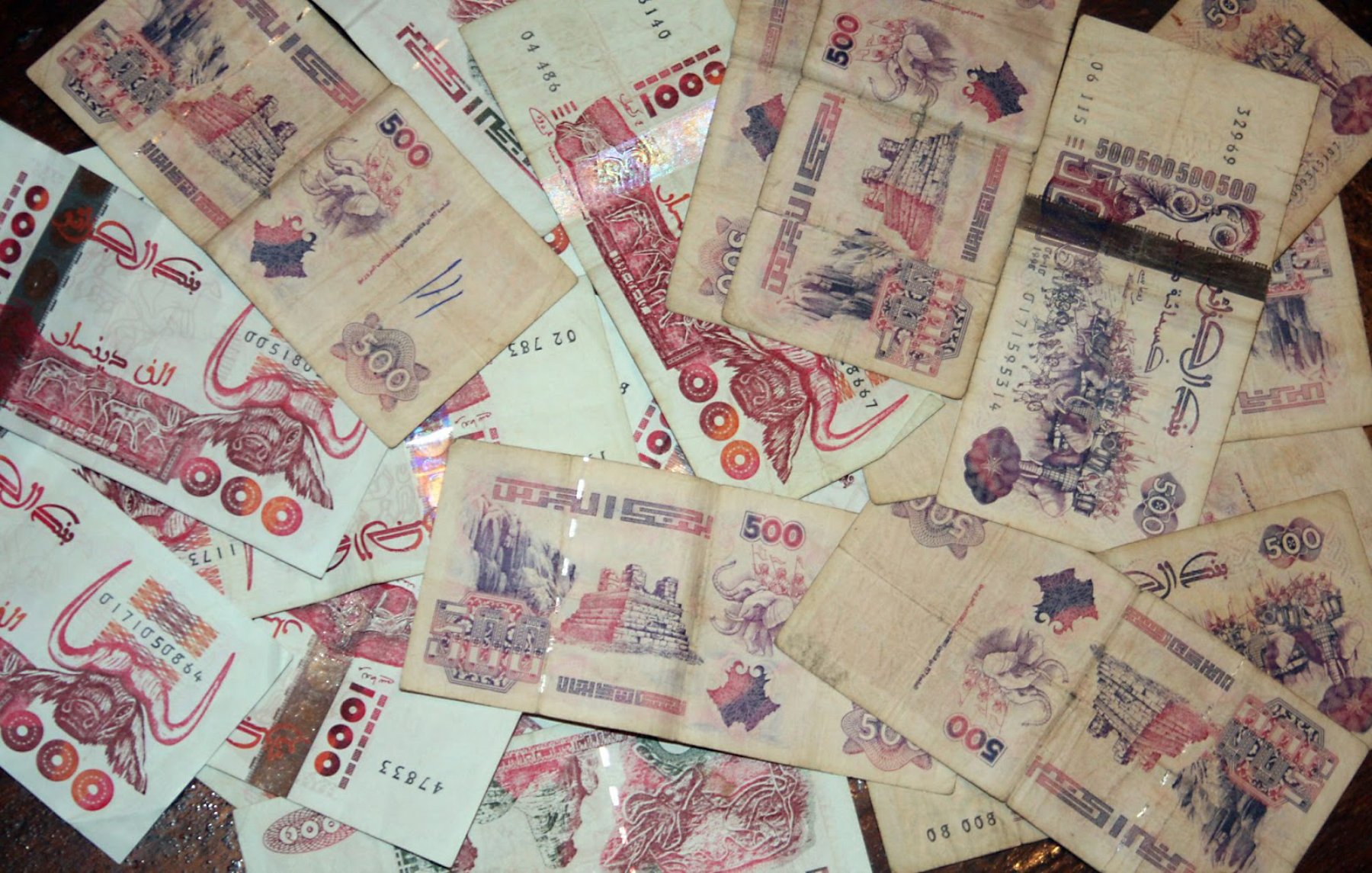 L'image montre un tas de billets de banque éparpillés, principalement de couleur beige avec des motifs et des textures variés. Certains billets sont de 500 unités, affichant des illustrations ou des inscriptions.
