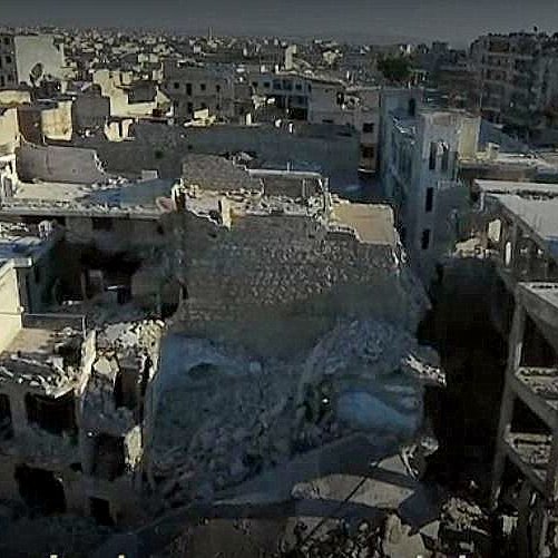 L'image montre un paysage urbain dévasté, probablement dans une zone de conflit. On y voit des bâtiments en ruines, certains complètement effondrés, d'autres partiellement intacts. Le sol est jonché de débris, témoignant des destructions causées par des bombardements ou des combats. L'environnement paraît déserté, avec peu ou pas de signes de vie, illustrant la tragédie et les conséquences dramatiques de la guerre sur les infrastructures urbaines.