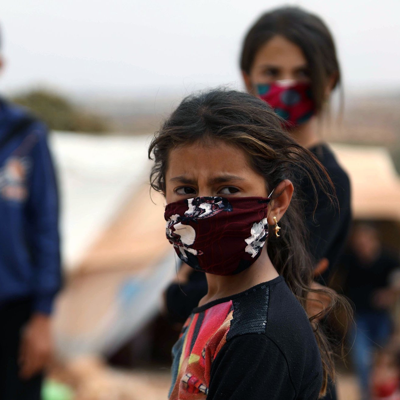 الصورة تظهر مجموعة من الأطفال في مخيم، حيث يرتدي بعضهم أقنعة واقية. في المقدمة توجد فتاة صغيرة تبدو قلقة، بينما خلفها يعبر الأطفال عن مشاعر متفاوتة. الخلفية غير واضحة، مما يشير إلى أنهم في بيئة مفتوحة لكنها غير مريحة. الإضاءة تبدو طبيعية، مما يعكس واقع الحياة اليومية في المخيم.