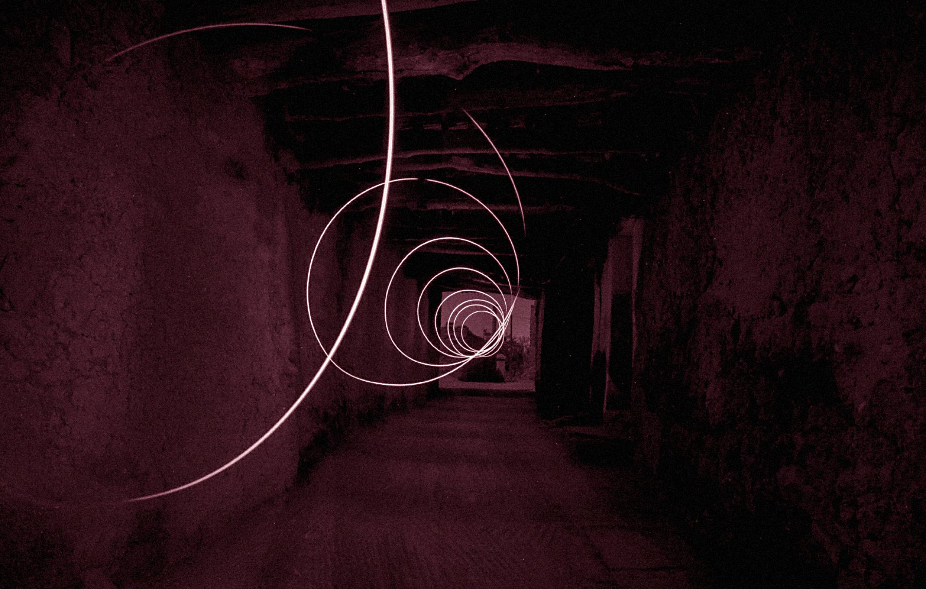 The image depicts a long, seemingly dark corridor with a series of swirling light trails creating a spiral effect. The color tone is predominantly red, contributing to a mysterious and somewhat surreal atmosphere. The walls of the corridor appear rough and textured, enhancing the sense of depth and perspective as the light trails guide the eye towards the far end of the hallway. The overall composition evokes a feeling of motion and intrigue.