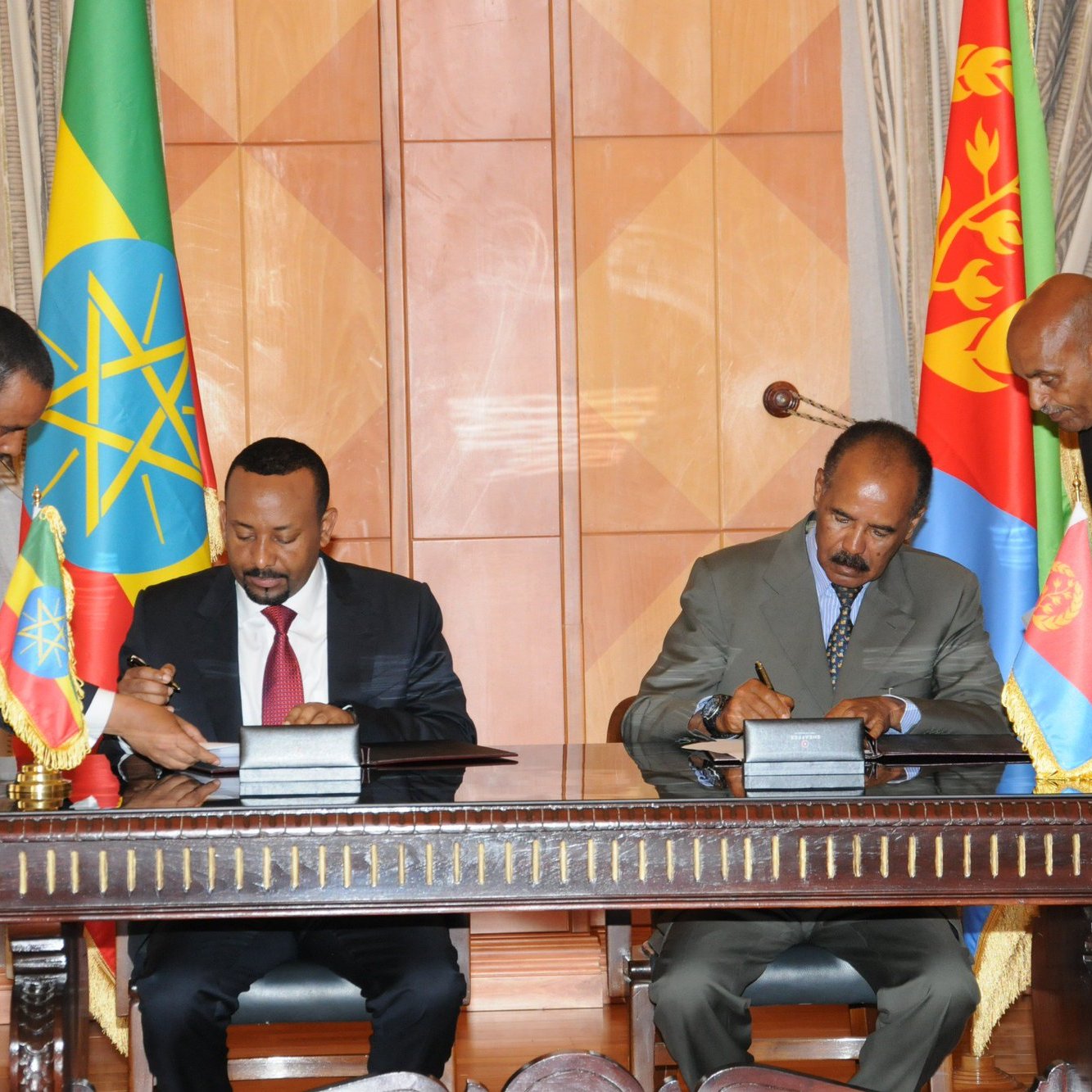 L'image montre une cérémonie de signature, probablement entre des représentants de l'Éthiopie et de l'Érythrée. Deux hommes, habillés en costume, signent des documents sur une table. À côté d'eux, des drapeaux éthiopien et érythréen sont visibles, ajoutant un élément symbolique à la scène. Les hommes semblent engagés dans un moment formel d'accord ou de collaboration.