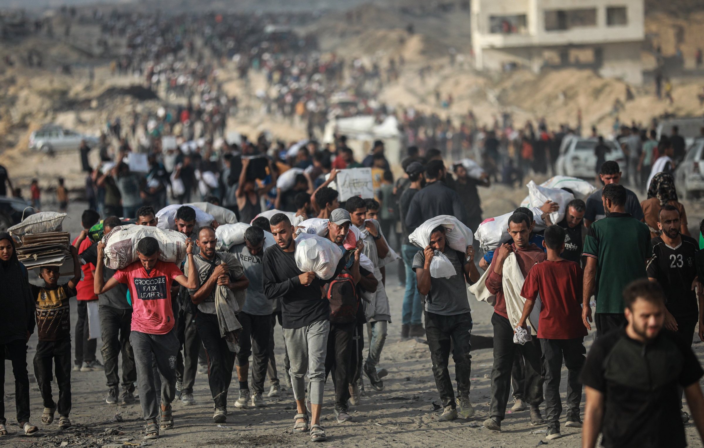 Gruppo di persone che trasportano sacchi su un terreno polveroso, in un contesto affollato.