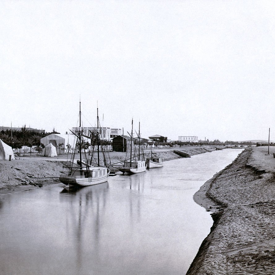 L'image montre une scène tranquille et nostalgique d'un canal ou d'une rivière. On y voit plusieurs bateaux amarrés, leur reflet se mêlant à l'eau calme. En arrière-plan, il y a des tentes et des structures qui évoquent une installation temporaire ou un camp. Le paysage environnant semble naturel, avec de la terre et des arbres. Cette photographie en noir et blanc donne une impression de sérénité et évoque une époque passée.