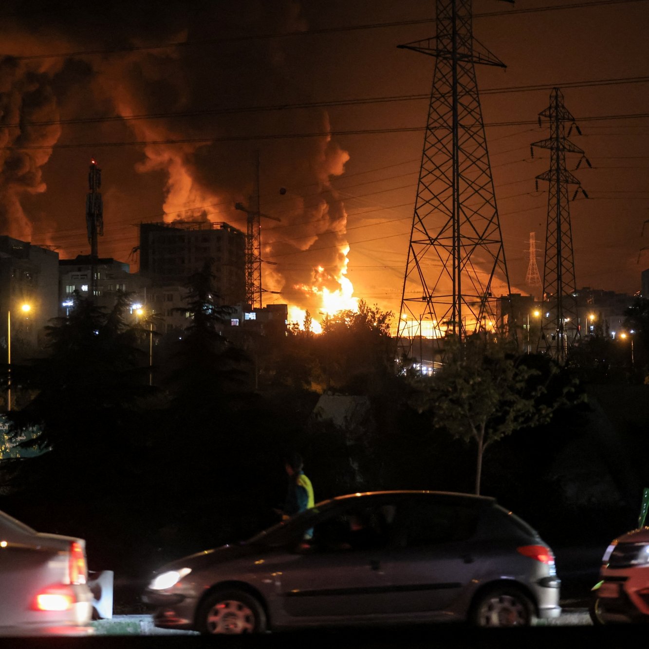 Un forte incendio illumina il cielo notturno, con fumi neri e auto in movimento.